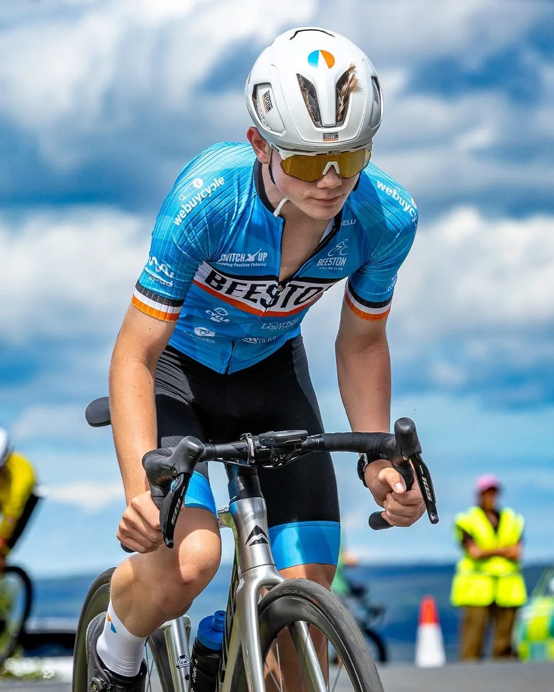 A cyclist wearing a white helmet, yellow-tinted sunglasses, and a blue jersey with 'BEESTON' written on it, riding a racing bicycle during a race, with spectators and officials in high-visibility vests in the background under a partly cloudy sky.