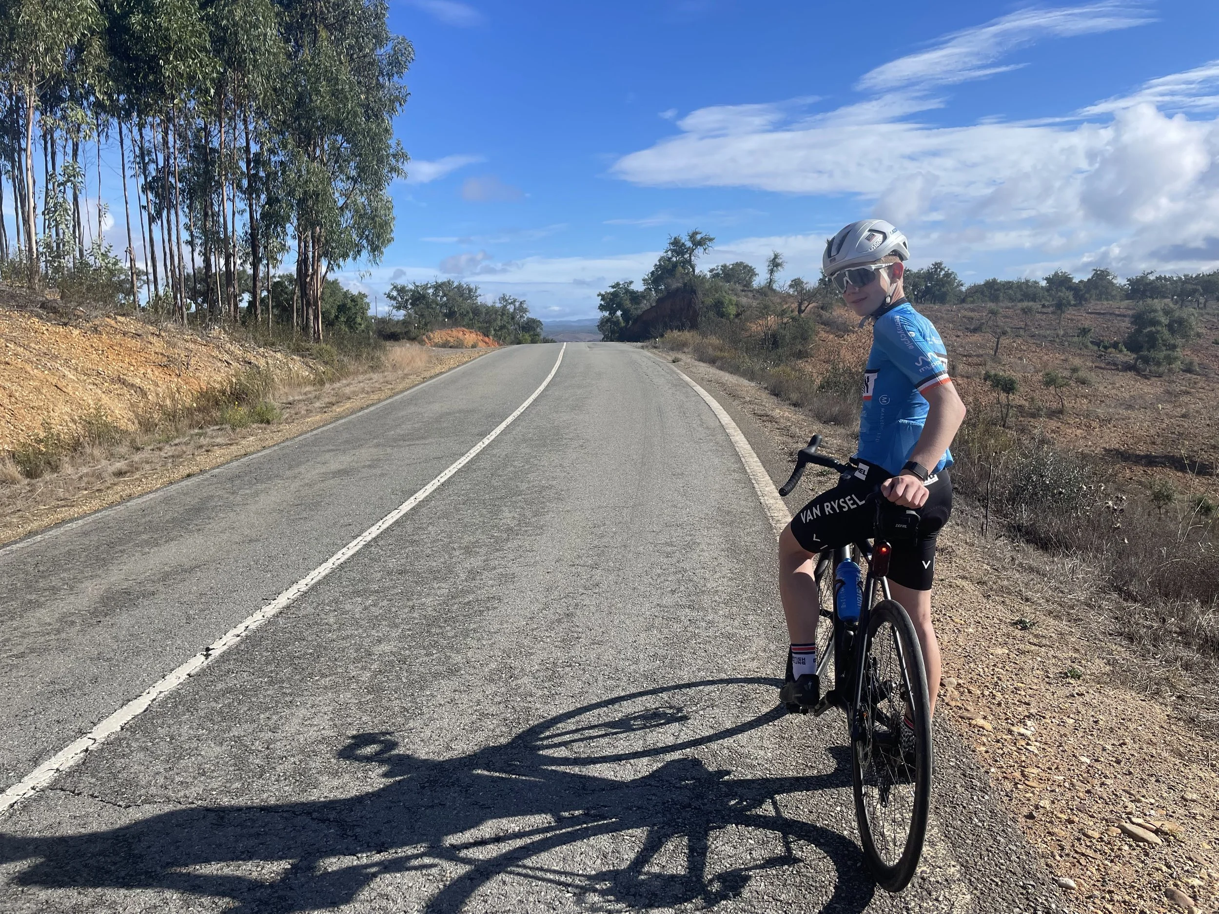 A person wearing a blue cycling jersey, black shorts, helmet, and sunglasses, riding a bicycle on a rural road with trees and open land on either side under a partly cloudy blue sky.