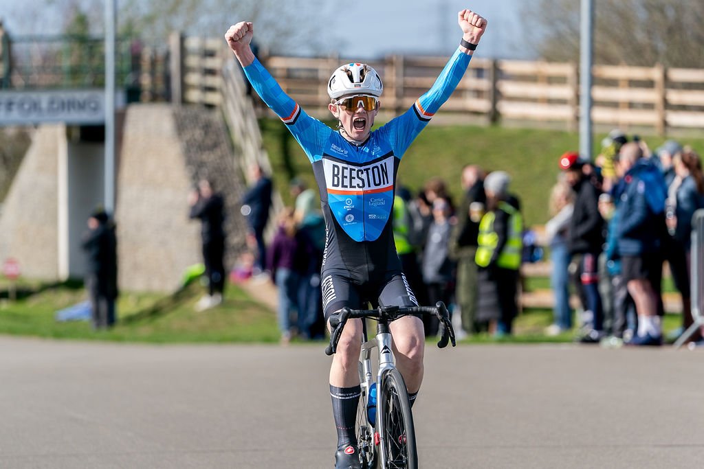 Cyclist crossing the finish line with arms raised in victory, wearing a blue and black uniform with a helmet, on a road with spectators in the background.
