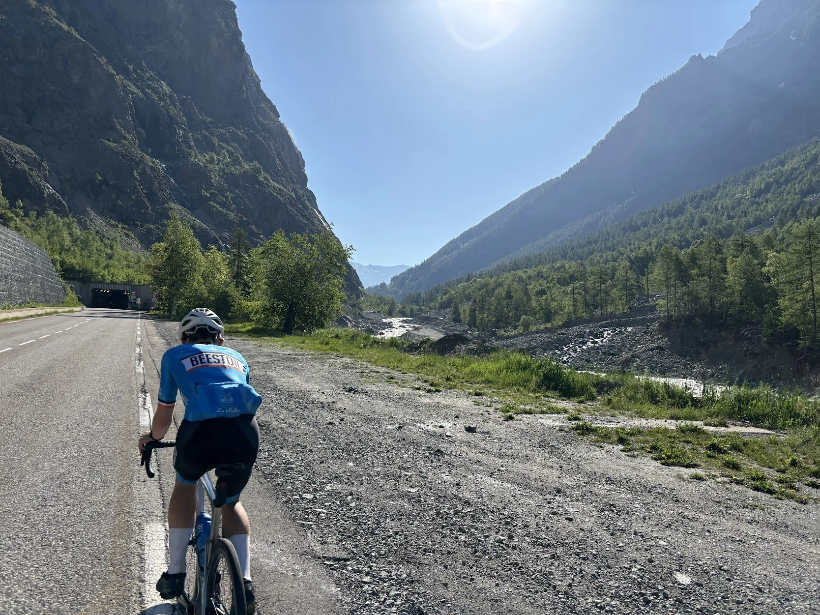 Cyclist riding on a mountain road through a scenic valley with green forests, rocky cliffs, and a stream.