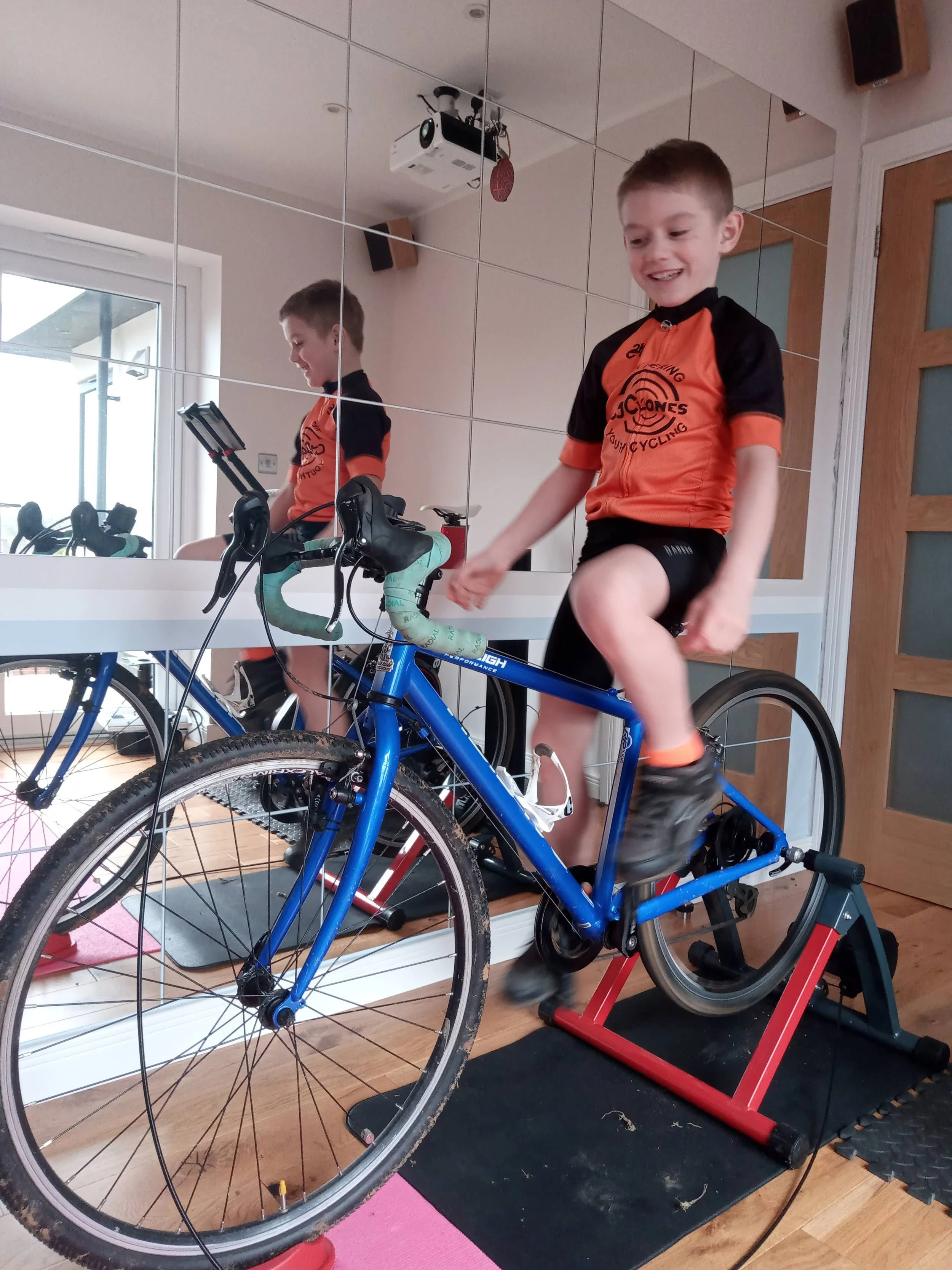 A boy on a blue bicycle inside a room, mounted on a trainer for indoor cycling, next to a mirror reflecting his image and the room's interior.