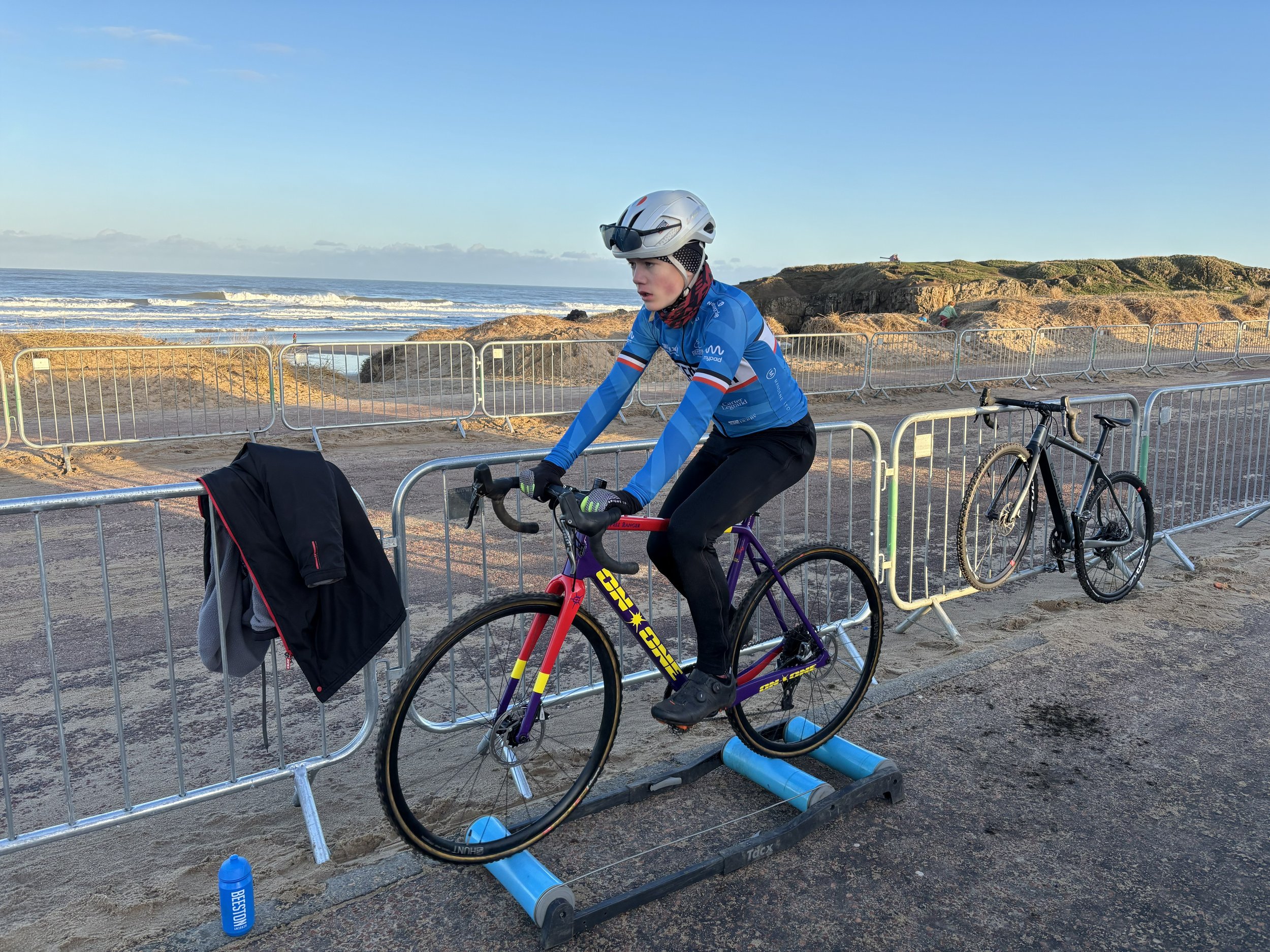 A person riding a bicycle along a beach with a metal barricade separating the path from the sandy area. The person wears a white helmet with sunglasses, a blue cycling jersey, and black pants. Another bicycle is parked nearby. The beach features rocks, sand, and ocean waves in the background under a clear sky.