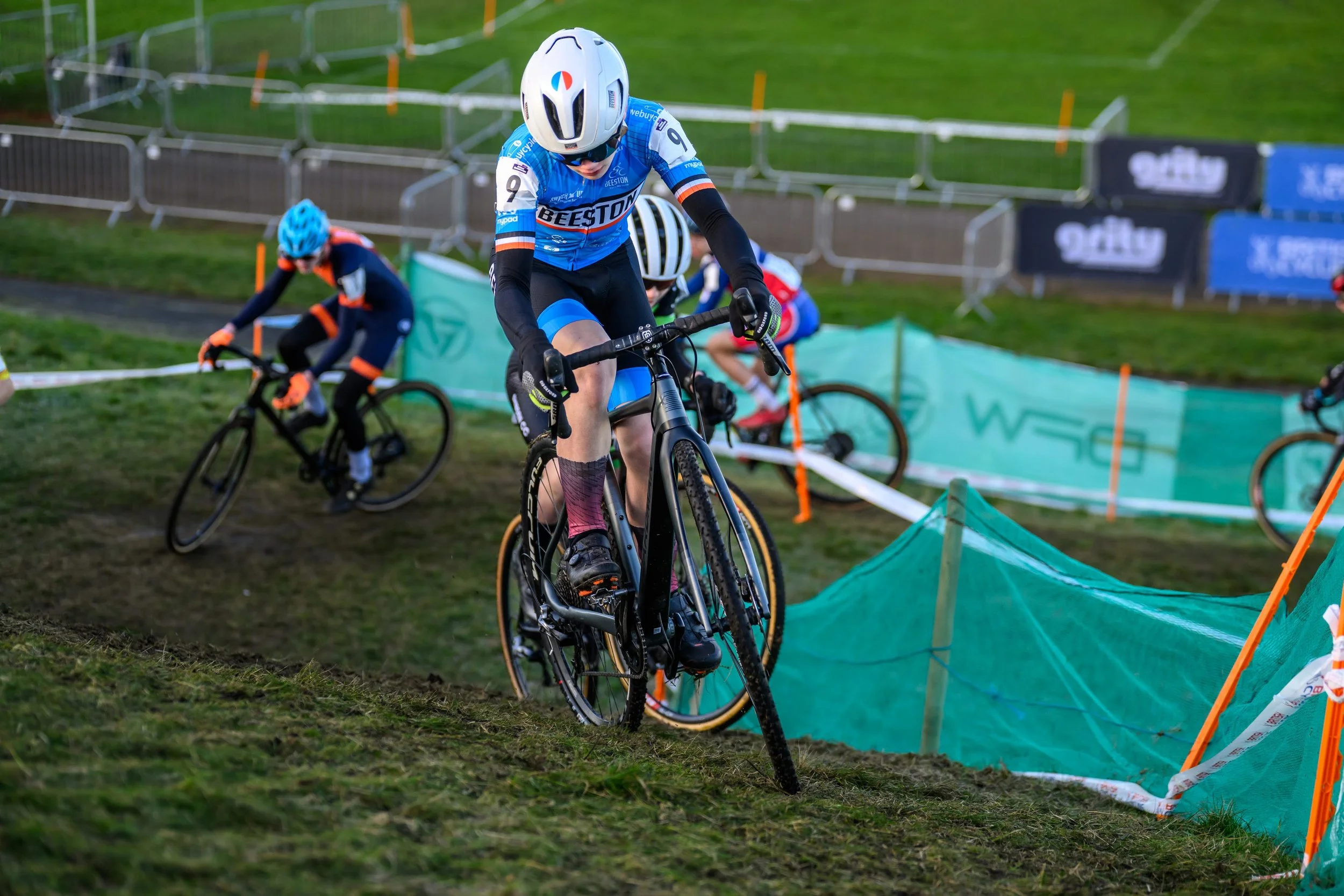 Cycling race with participants navigating through a marked off-road section, wearing helmets and team jerseys, on a grassy course with fencing and banners.