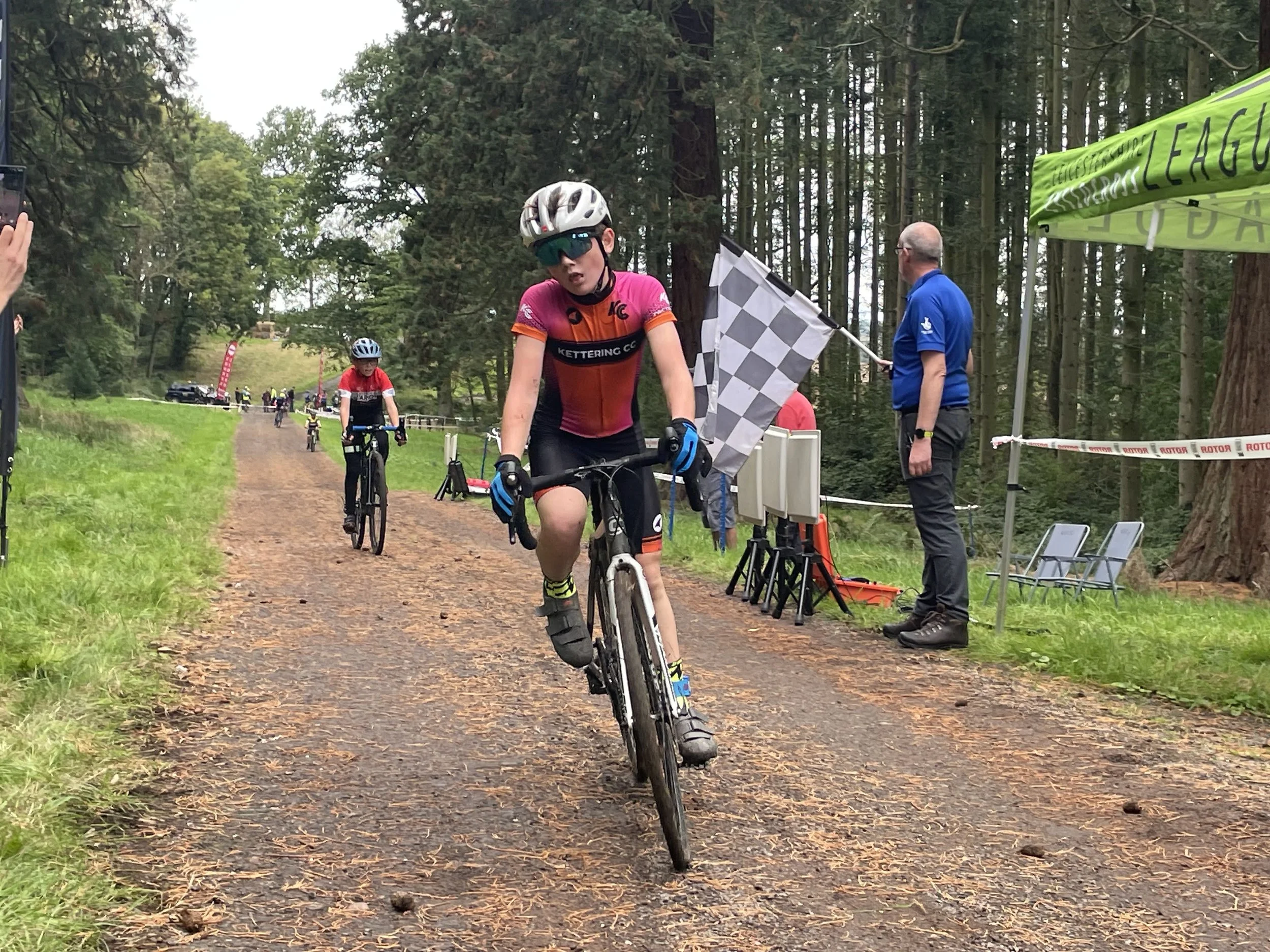 A young cyclist wearing a pink and orange jersey, a white helmet, and sunglasses finishes a mountain bike race in a wooded area. A man in a blue shirt is holding a checkered flag near the finish line, with a green tent and several other cyclists and spectators in the background.