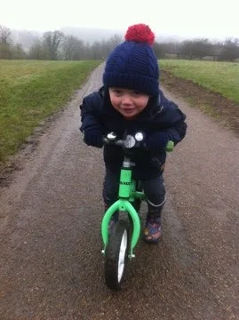 Young child riding a green balance bike on a dirt path, wearing a blue winter coat and a blue knit hat with a red pom-pom, in an outdoor rural setting.