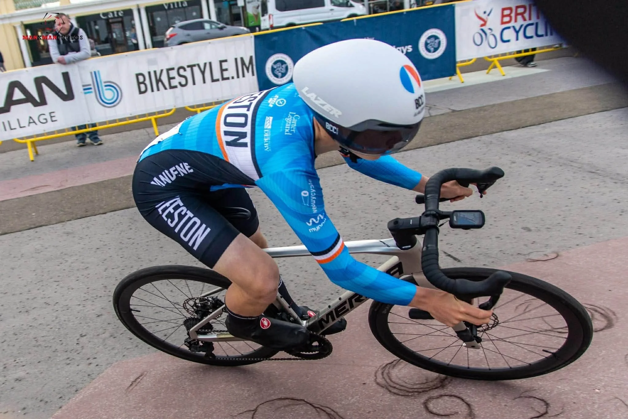 A cyclist in a blue racing outfit biking on a city street during a race, with spectators and banners behind.