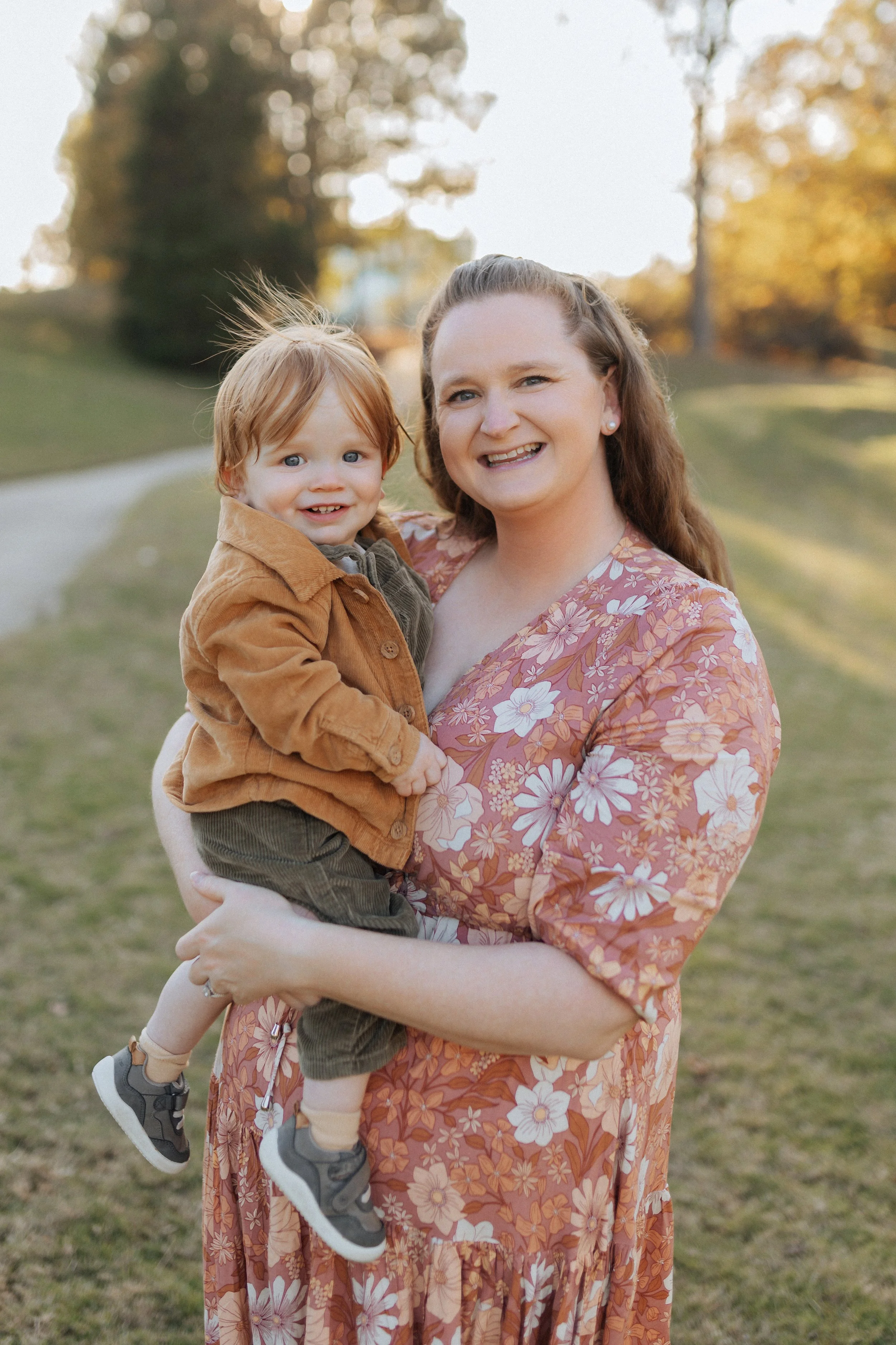 A woman with long, wavy brown hair wearing a floral dress holding a young boy with red hair in a brown jacket outdoors on a sunny day. The background features trees and a grassy area.