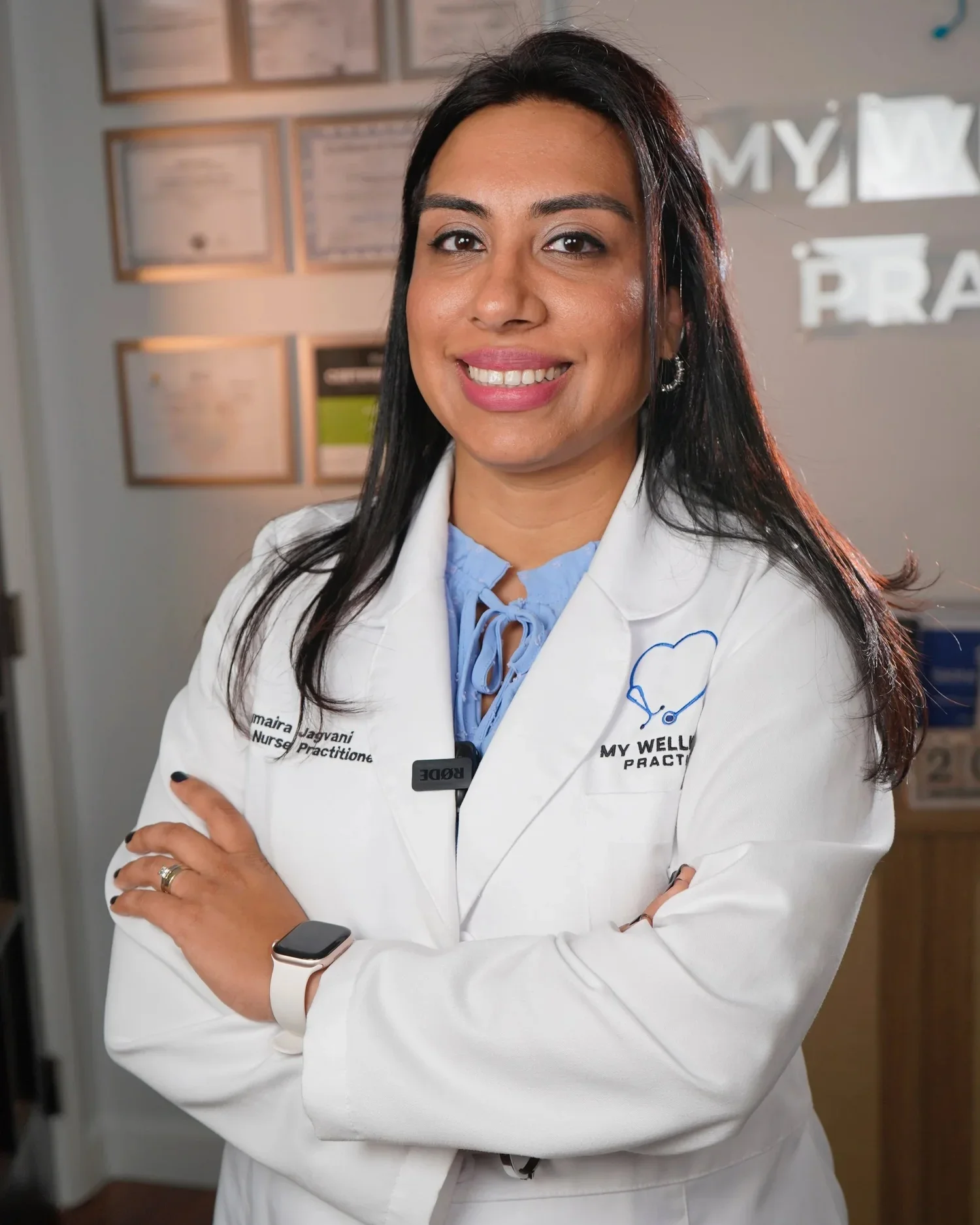 A smiling female nurse in a white coat standing with arms crossed in a medical office. She wears a smartwatch and has a stethoscope logo embroidered on her coat along with her name and title.