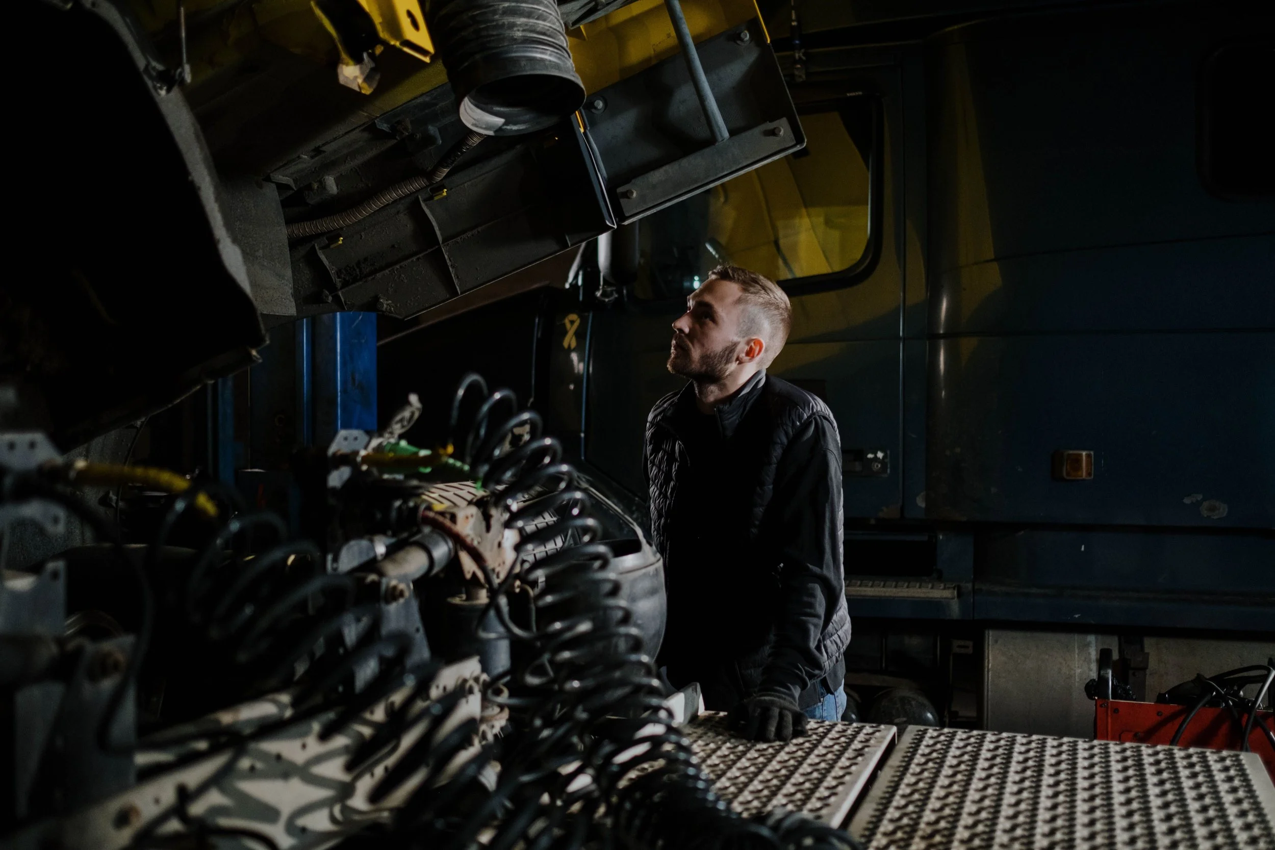 A man working on electronic components inside an industrial or repair shop, surrounded by cables, wires, and equipment with a yellow vehicle partially visible in the background.