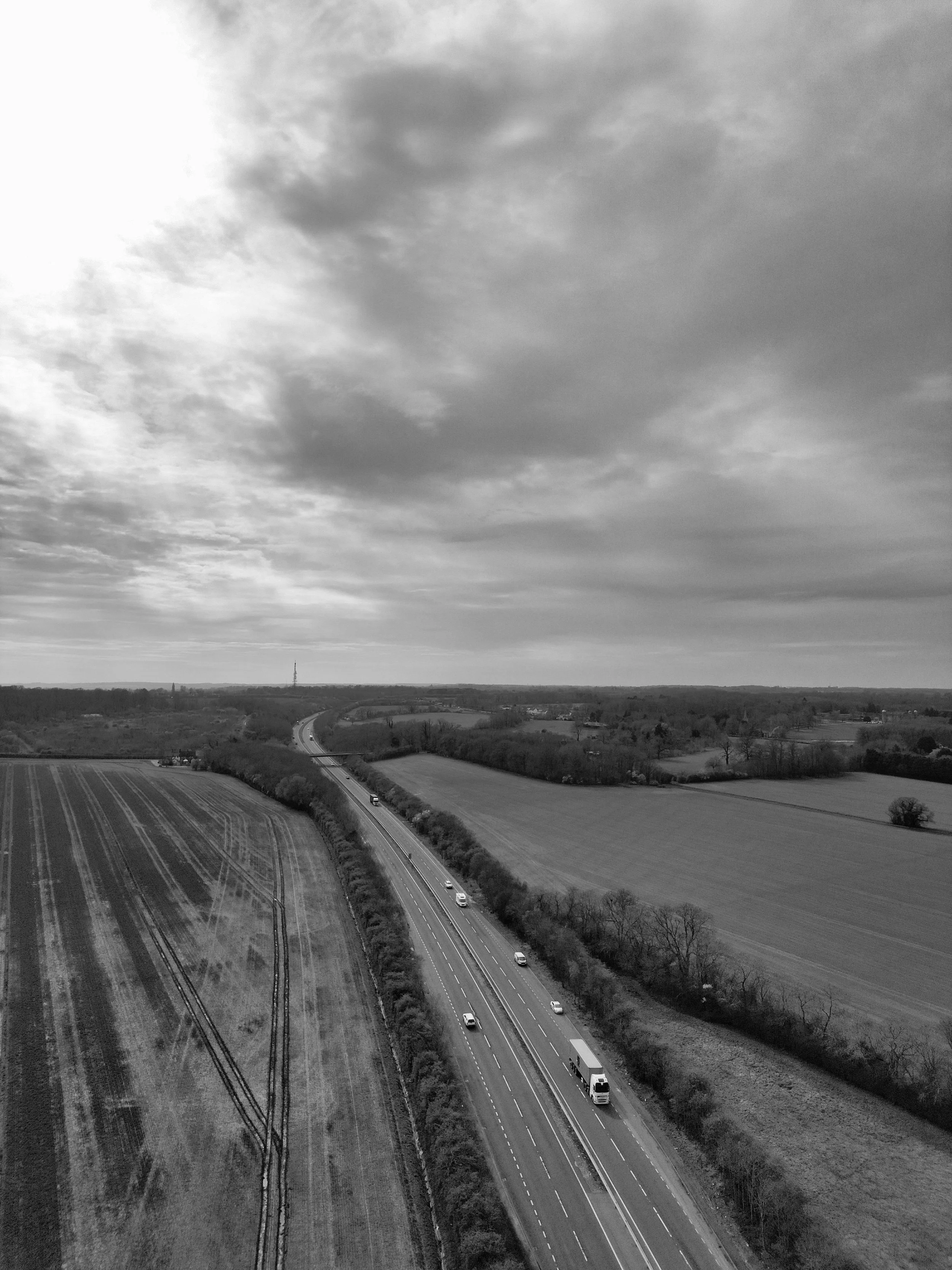 A black and white aerial view of a rural landscape with fields, a highway with vehicles, and a cloudy sky.
