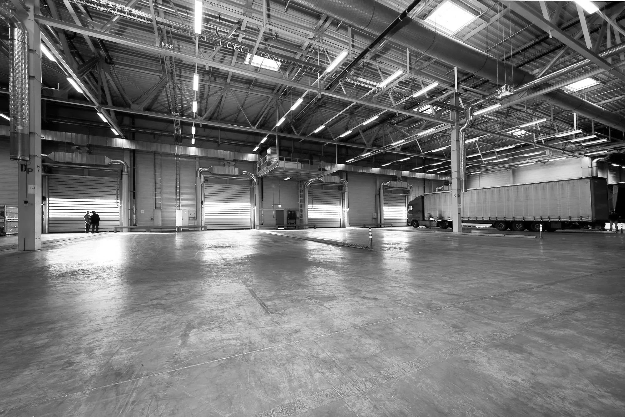 A large, empty warehouse or industrial space with a concrete floor, multiple large roll-up doors, and a truck parked inside. The ceiling has exposed beams, pipes, and fluorescent lighting, with a few small figures visible near the doors.