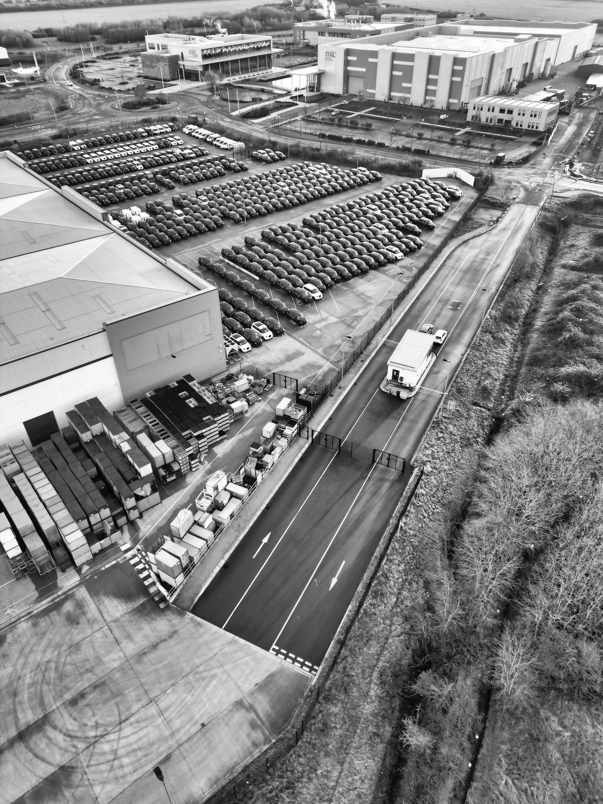 Aerial view of a large parking lot with numerous parked cars, adjacent to an industrial or commercial building with a curved road and surrounding landscape, shown in black and white.