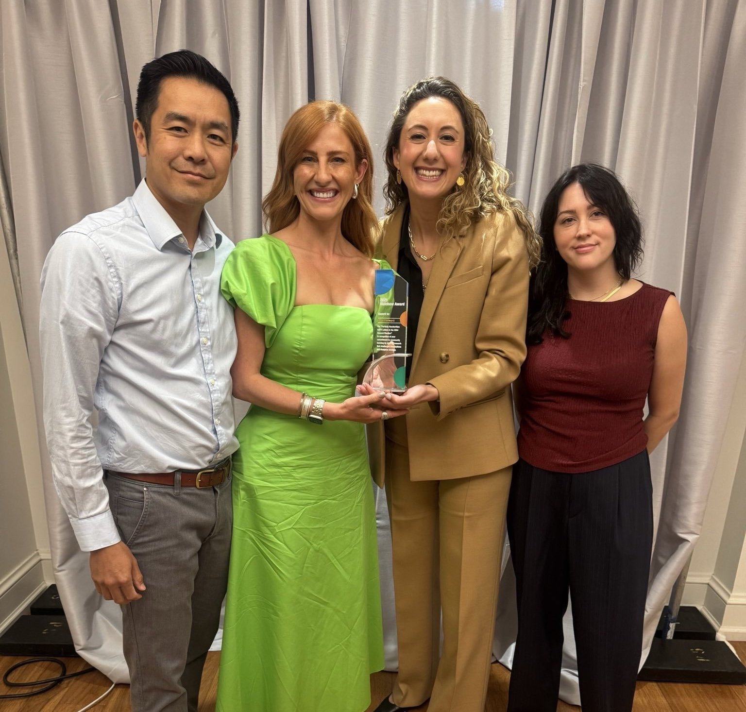 Four people standing together, smiling, with one woman holding a trophy or award in the center, in front of a curtain backdrop.