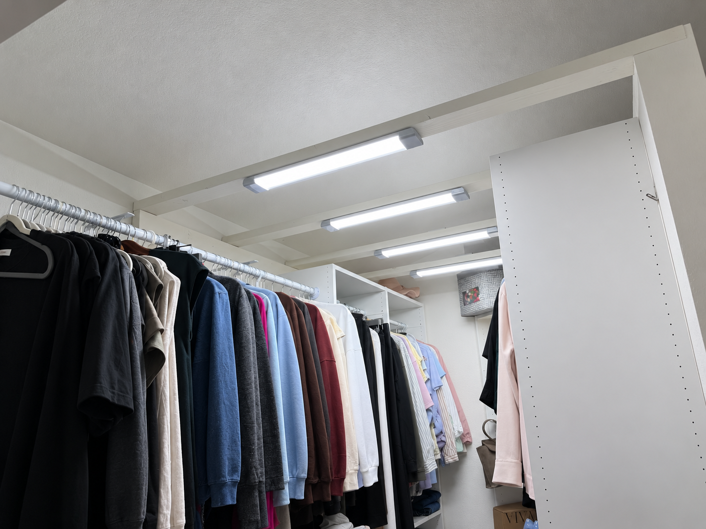 A closet with hanging clothes on a rack, including hoodies, shirts, and jackets, with shelves above and a tall white cabinet.