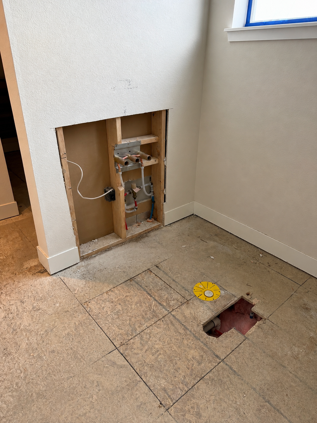Kitchen wall with plumbing and drywall removed near a window, exposed pipes and an unfinished floor with a small hole and a yellow plastic cover.