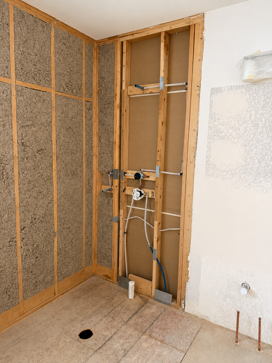 Bathroom wall under renovation showing exposed plumbing and electrical wiring, with insulation and drywall partially removed.