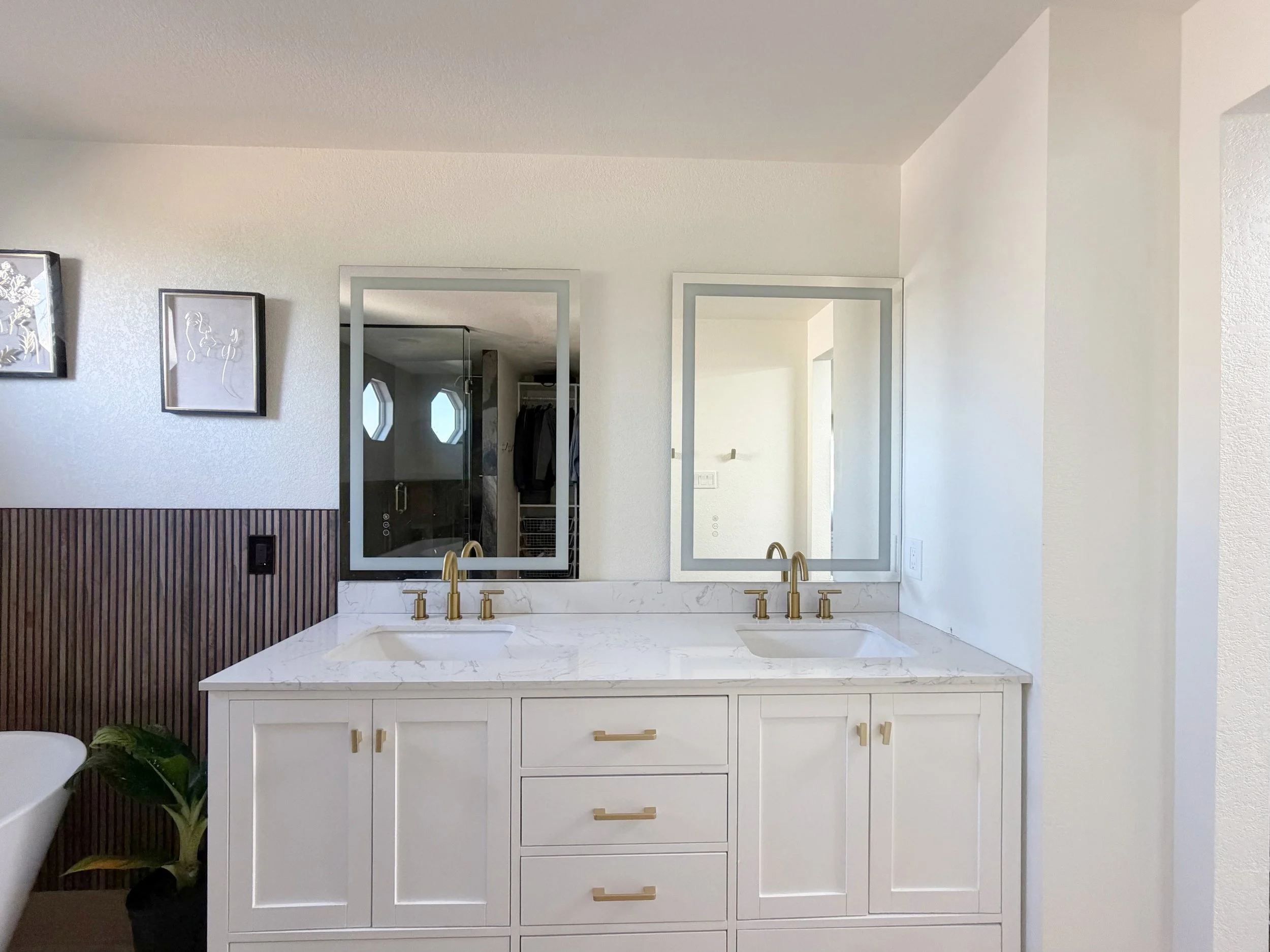White double vanity with gold faucets, two illuminated mirrors, and a wood accent wall in a completed Vexo Homes bathroom.