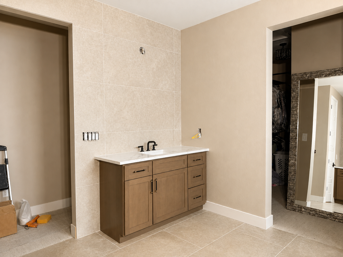 Bathroom vanity with a white marble countertop and black fixtures, beige tiled wall, and open closet with hanging clothes reflected in a large mirror.