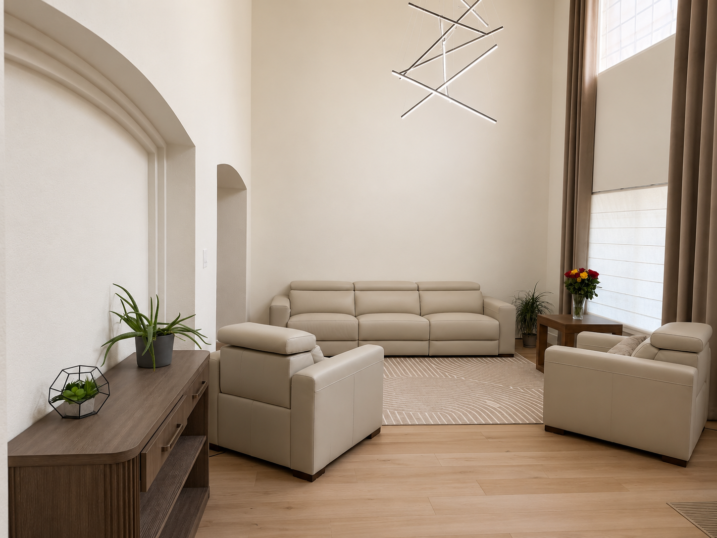 Modern living room with beige leather sofa, armchairs, wooden side table with flowers, and potted plants, illuminated by large window with curtains.