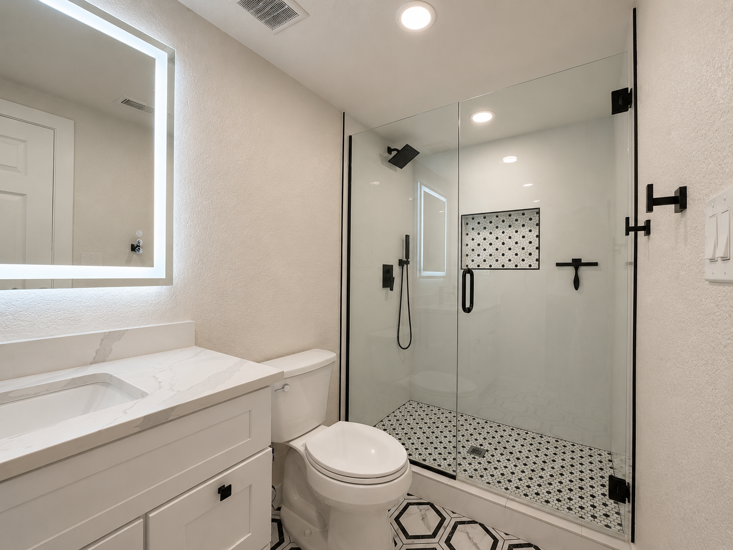 Modern bathroom with white vanity, toilet, and glass shower enclosure with black hardware, black and white mosaic tile shower floor, built-in niche, and geometric black and white patterned floor.