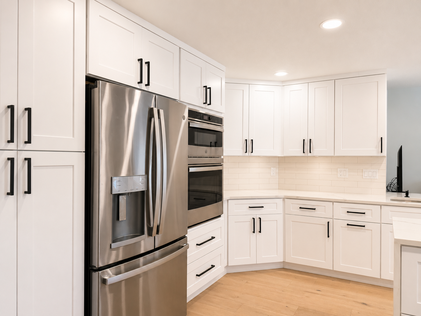 Modern kitchen with white cabinets, stainless steel refrigerator and double oven, light wood flooring, and white tiled backsplash.