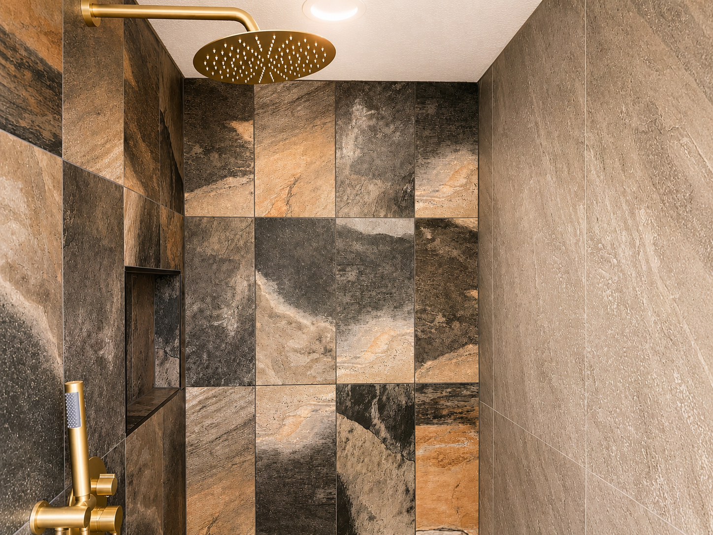 A shower with dark, multicolored stone tiles on the wall, a large round ceiling shower head, and a gold shower control panel with a handheld shower.