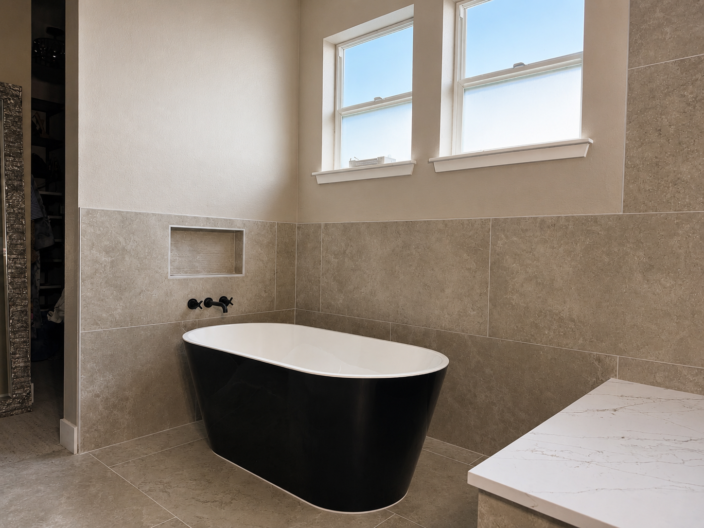 A modern bathroom with a black free-standing bathtub, two high small windows, beige textured tiled walls, and a built-in niche for toiletries.