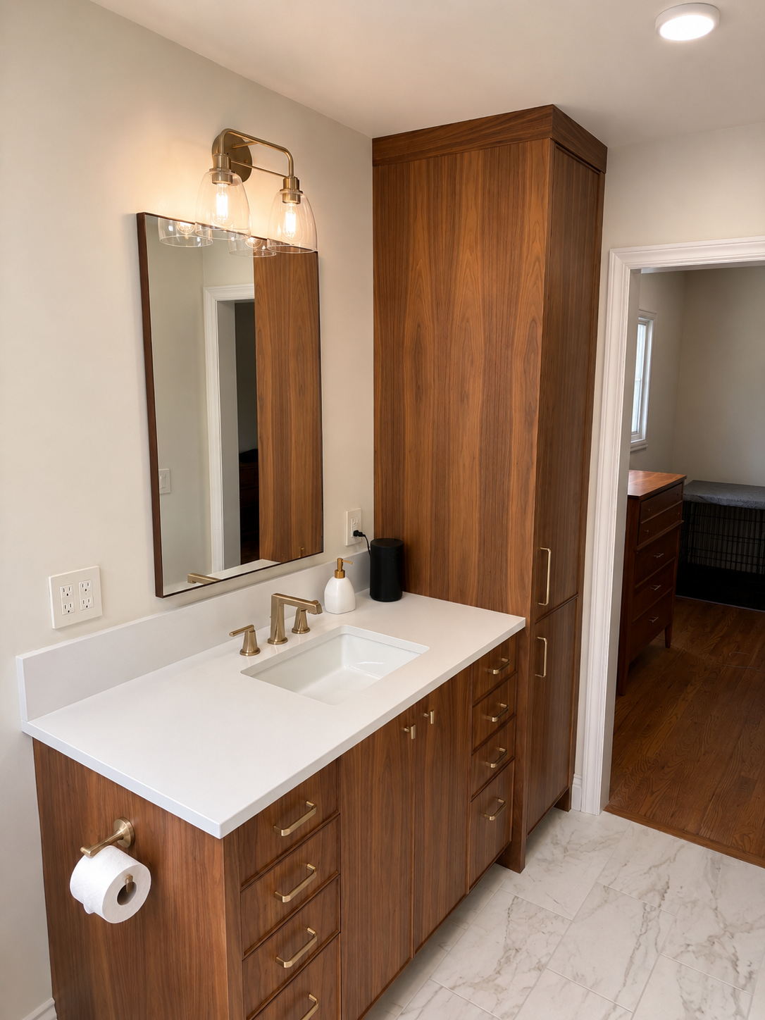 Bathroom vanity with wooden cabinets, white countertop, rectangular undermount sink, brass faucet, mirror, wall-mounted light fixture, black speaker, soap dispenser, toilet paper holder, and door leading to another room with a dresser and window.