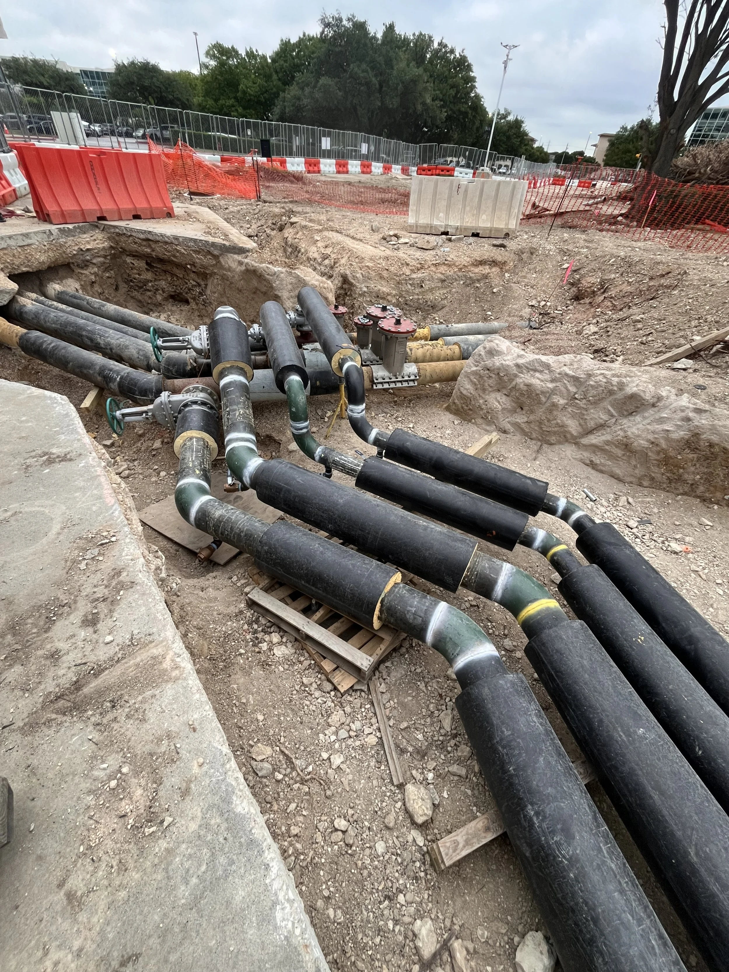 Construction site with large black pipes laid in a trench, surrounded by dirt and rocks, with orange safety barriers and fencing in the background.
