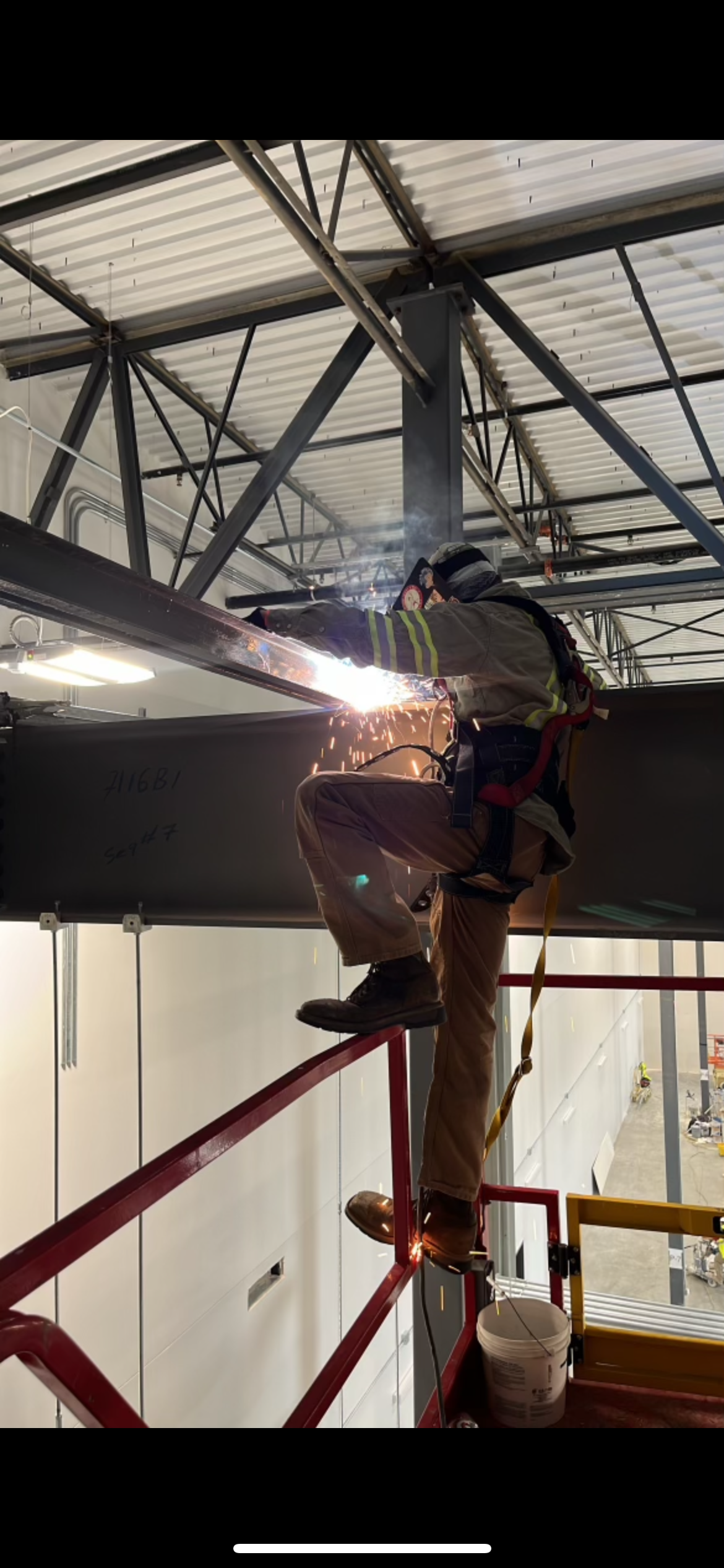 A worker welding a large metal pipe inside an industrial building, wearing safety gear and standing on a scissor lift.
