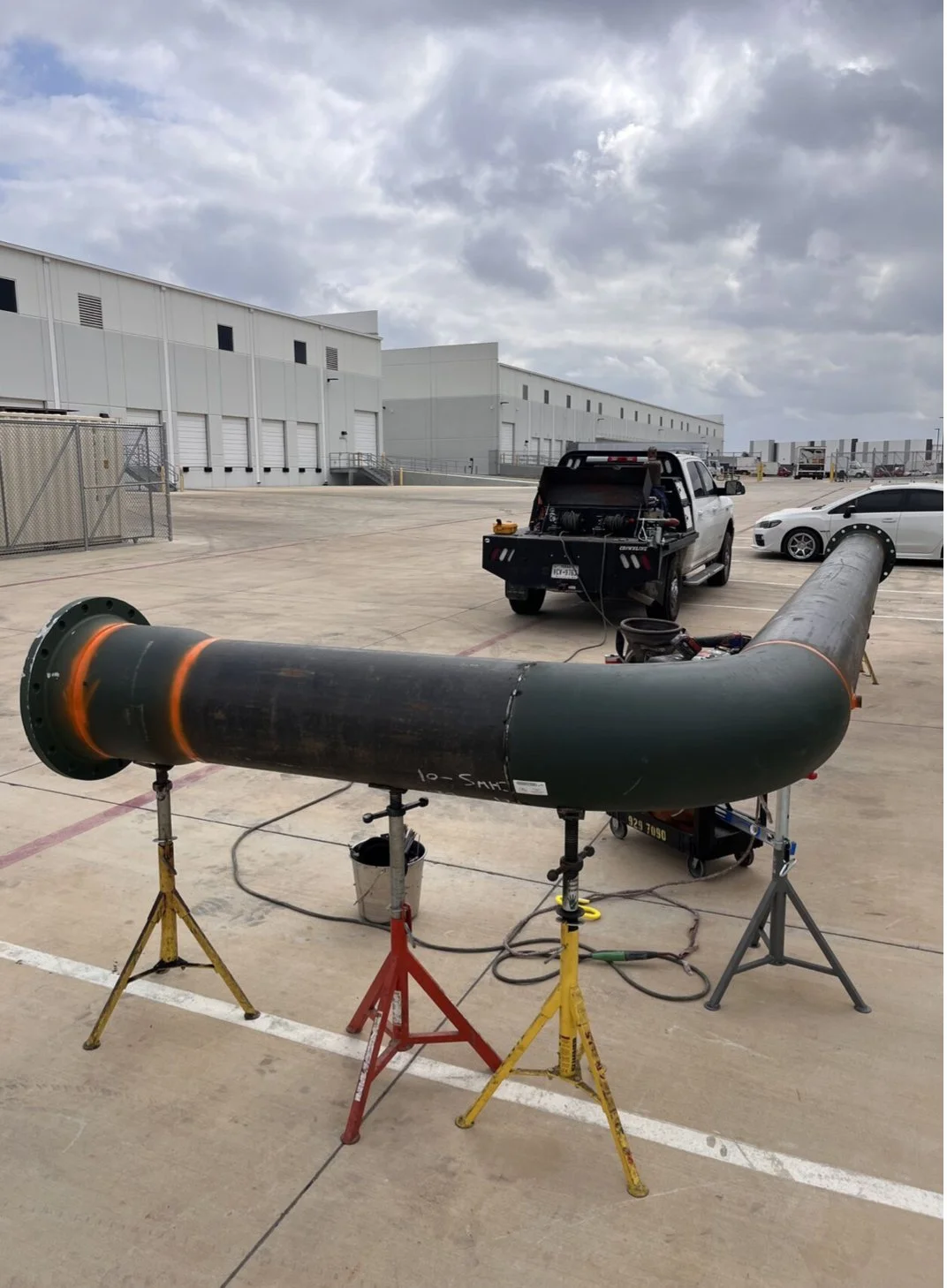 Large black pipe on metal stands with a connected device and a bucket, set on a concrete surface outside a warehouse with vehicles and cloudy sky in the background.
