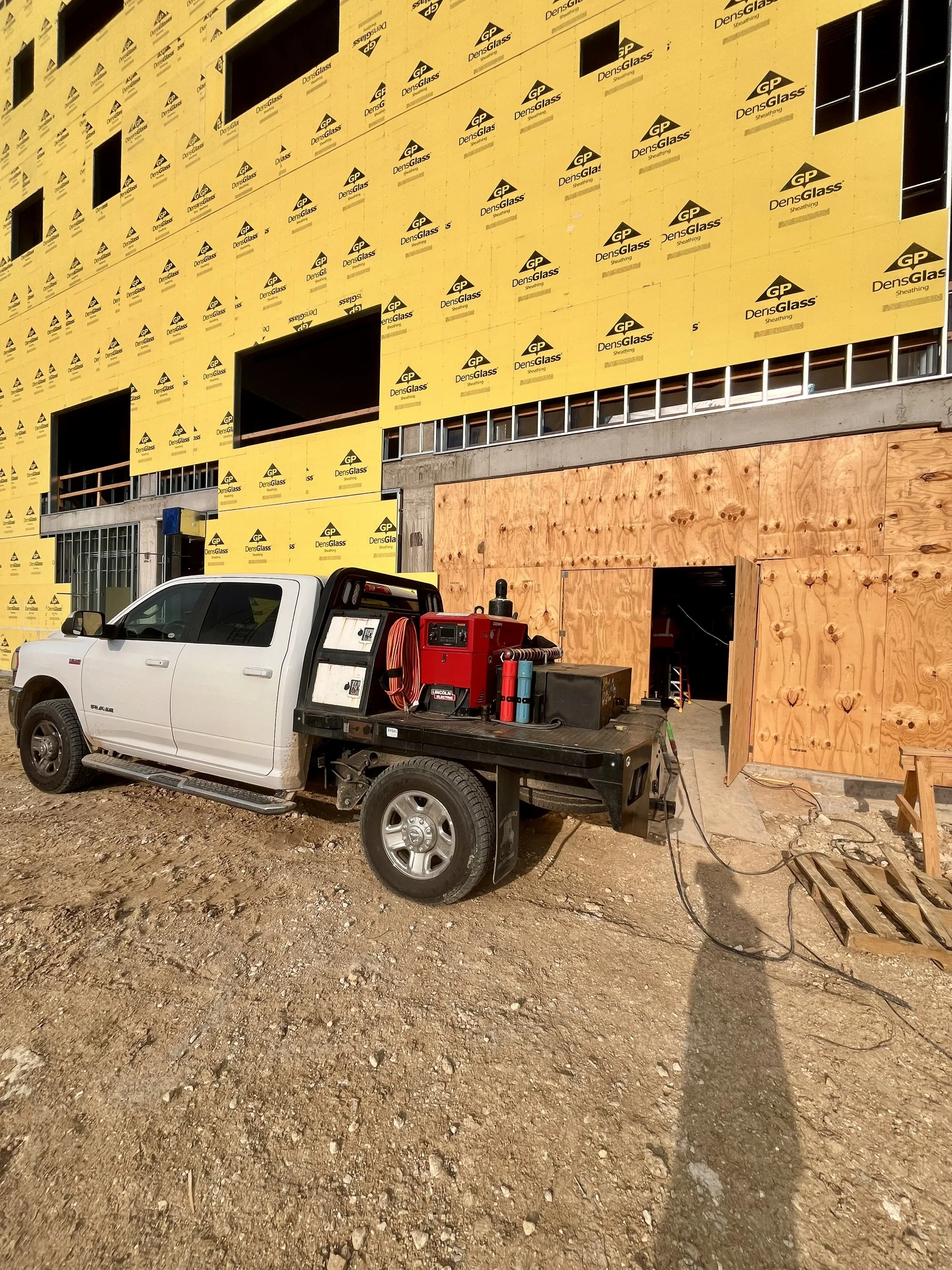 Construction site with a partially built building covered in yellow DensGlass sheathing, showing window openings, with a white pickup truck and construction equipment parked on dirt ground in front.