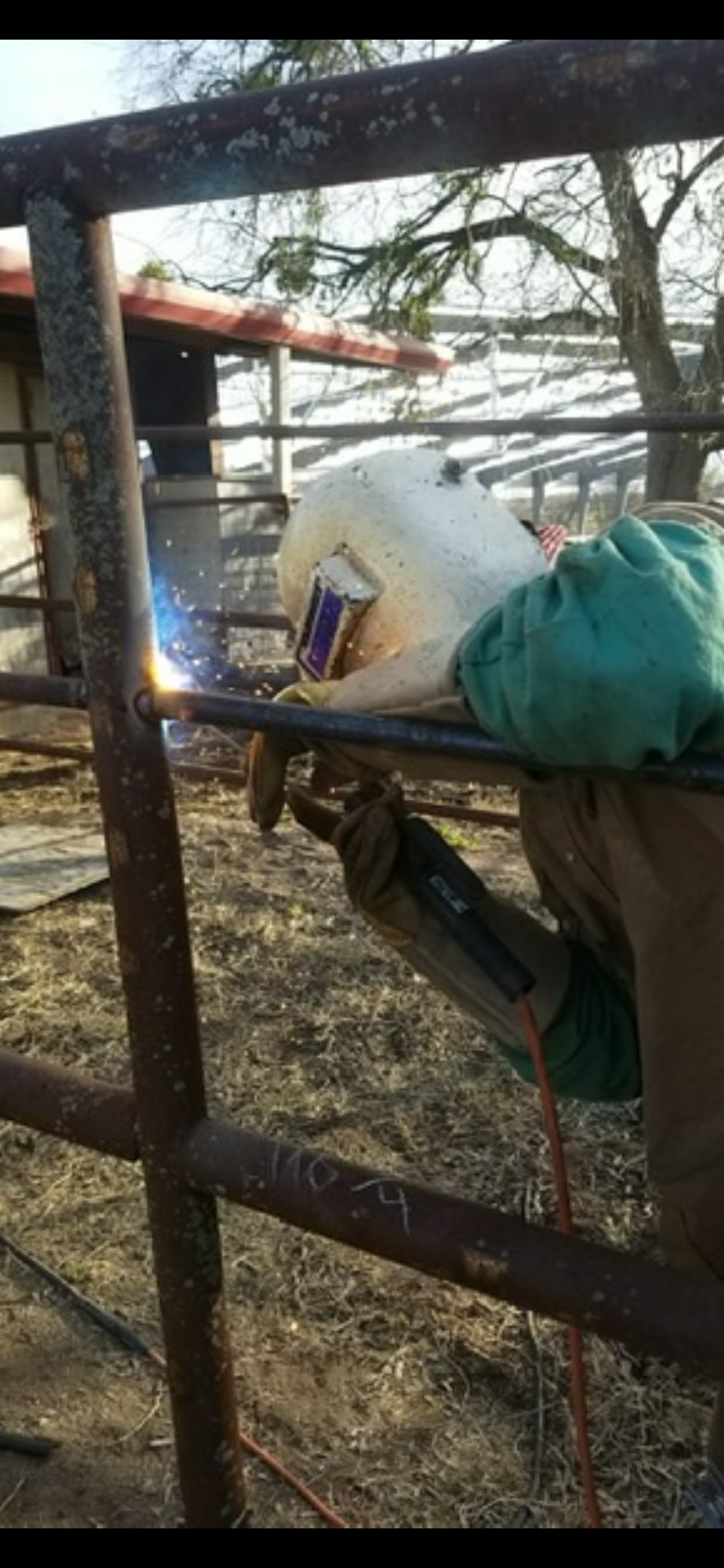 Person welding on a rusty metal structure outdoors, with trees and a building in the background.