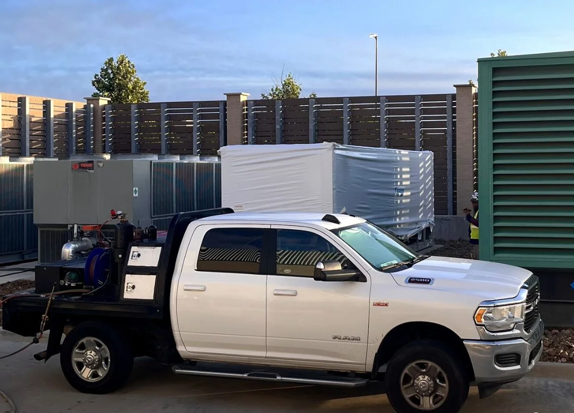 White pickup truck with industrial equipment in the bed, parked outdoors near a green vent, with a fence and small trees in the background.