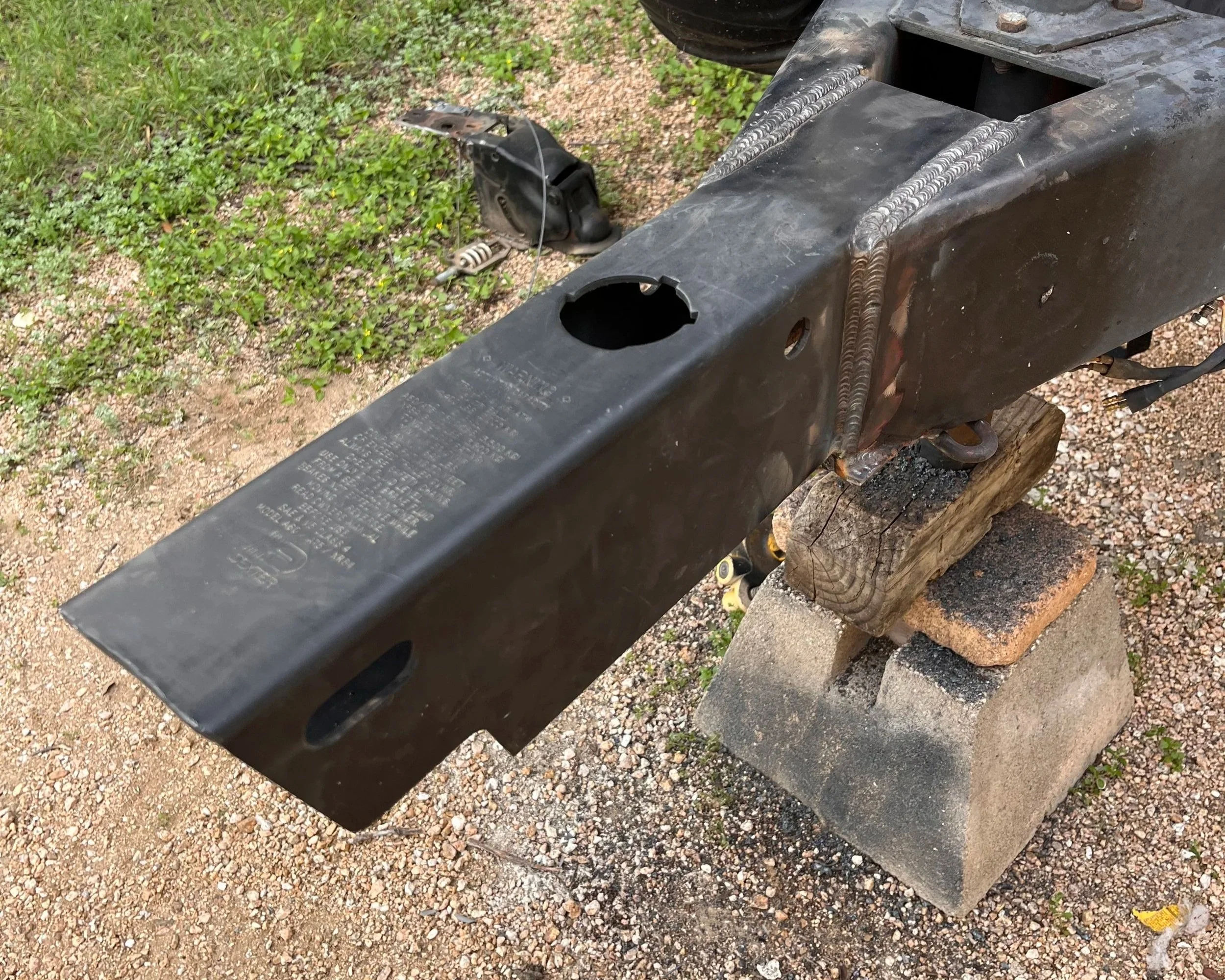 A black metal trailer frame with a circular cutout on top, resting on a wooden and concrete support on gravel ground, with some grass and bushes in the background.