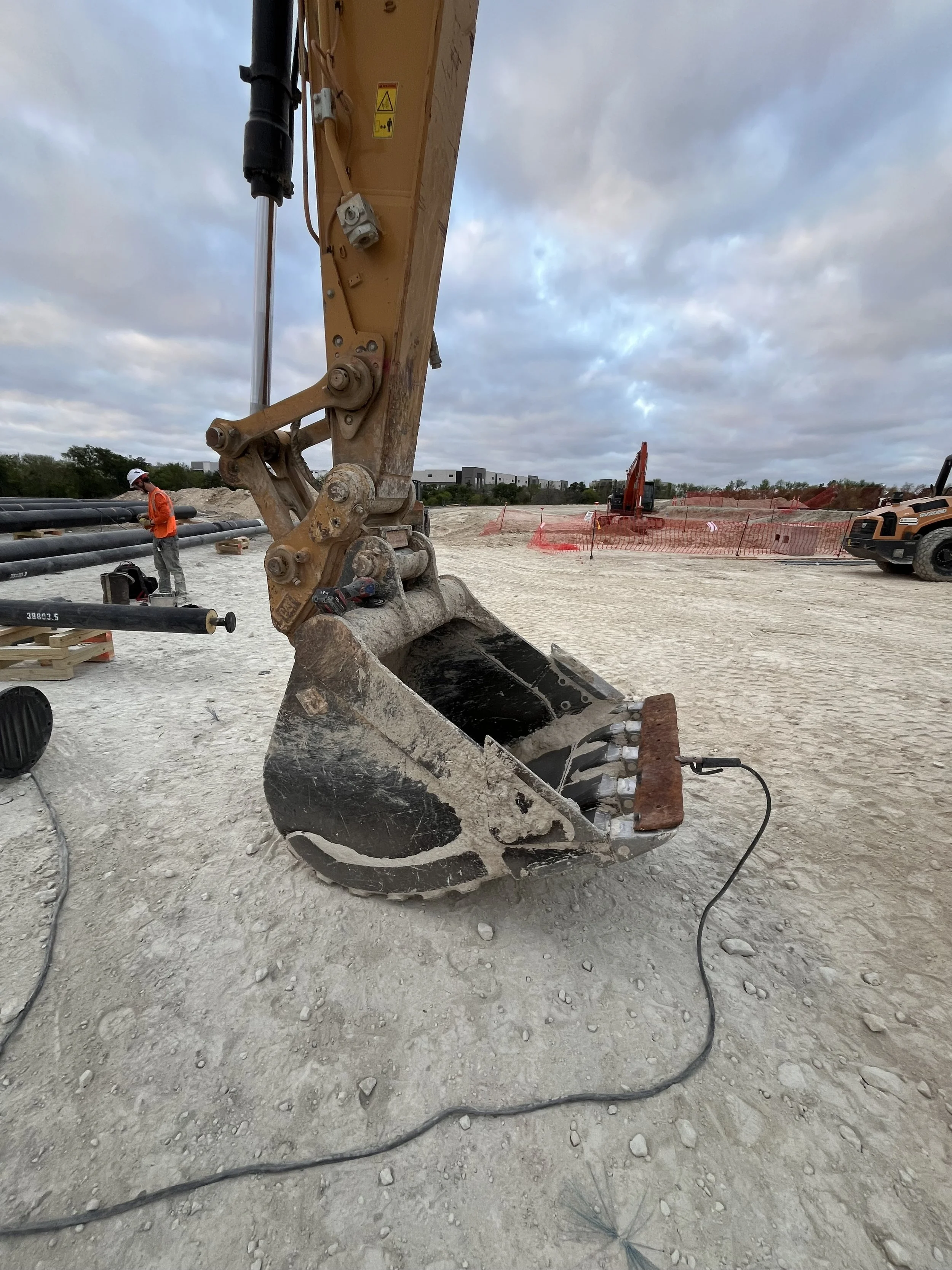 Construction site with an excavator bucket in the foreground, a worker in an orange safety vest and white helmet on the left, pipes laid out on the ground, and another excavator in the background, under a cloudy sky.