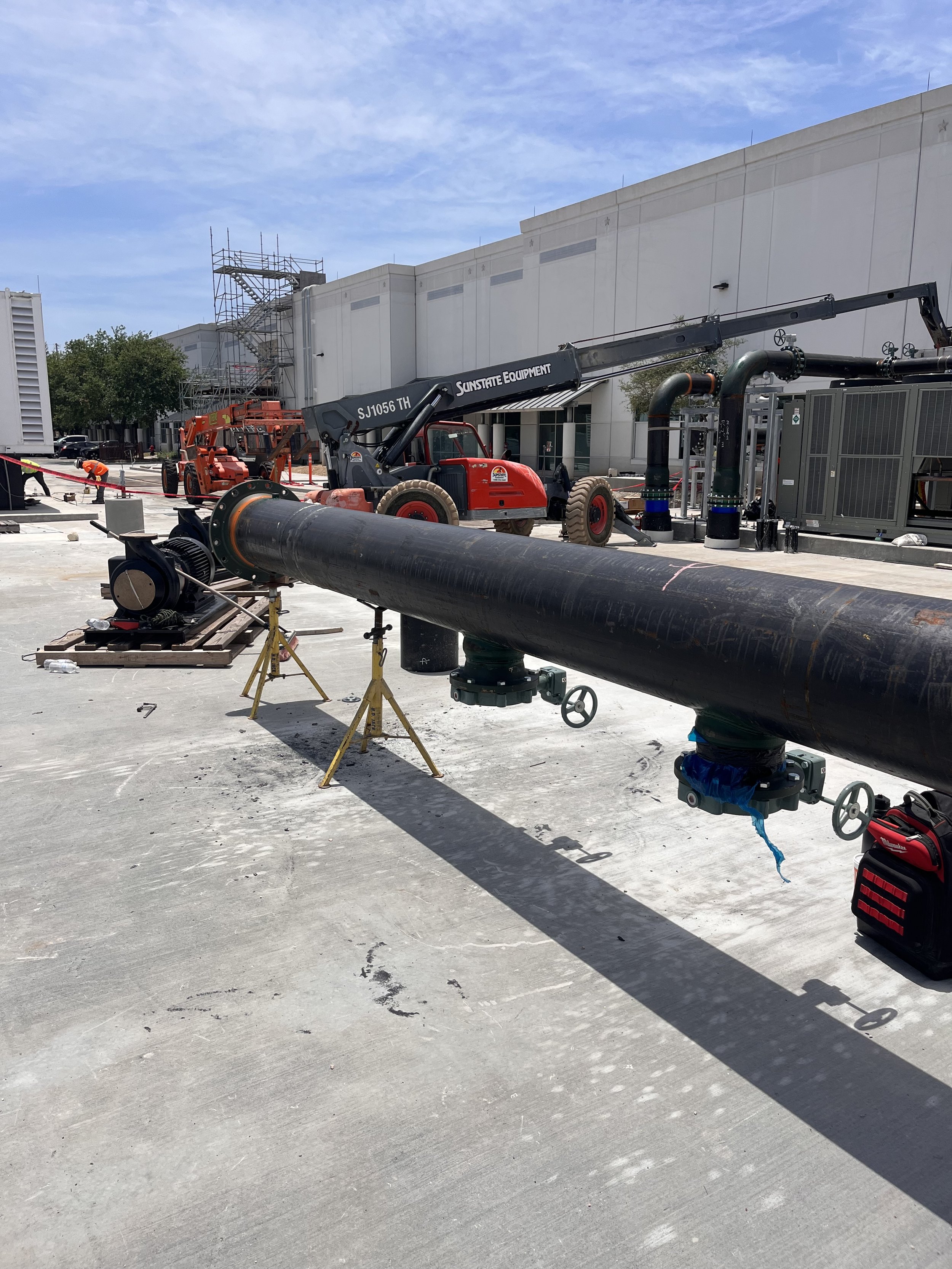 Construction site with large black pipeline, orange machinery, and a crane arm on a sunny day with a white building and blue sky in the background.