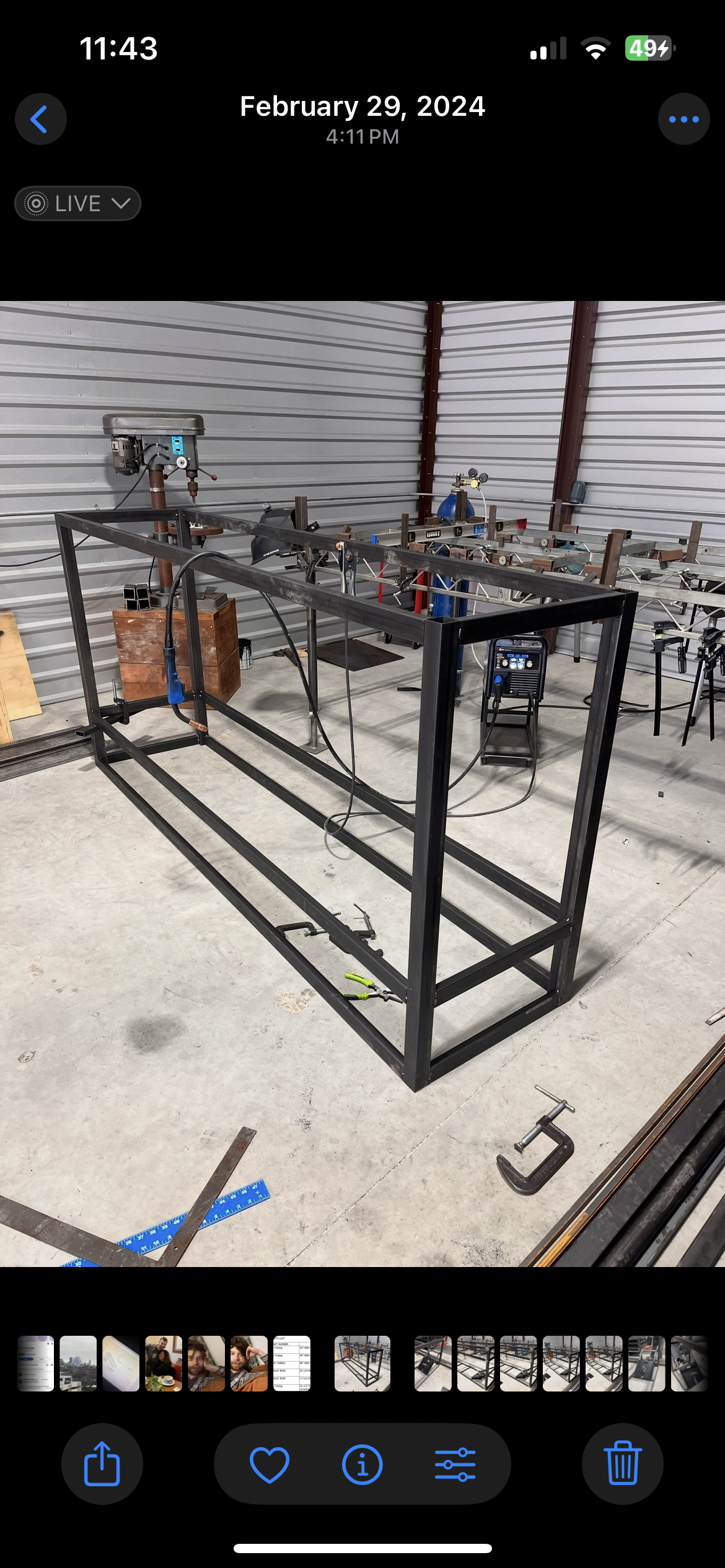 A workspace with a black metal frame structure, a drill press, and various tools and equipment on a concrete floor inside a metal-walled building.