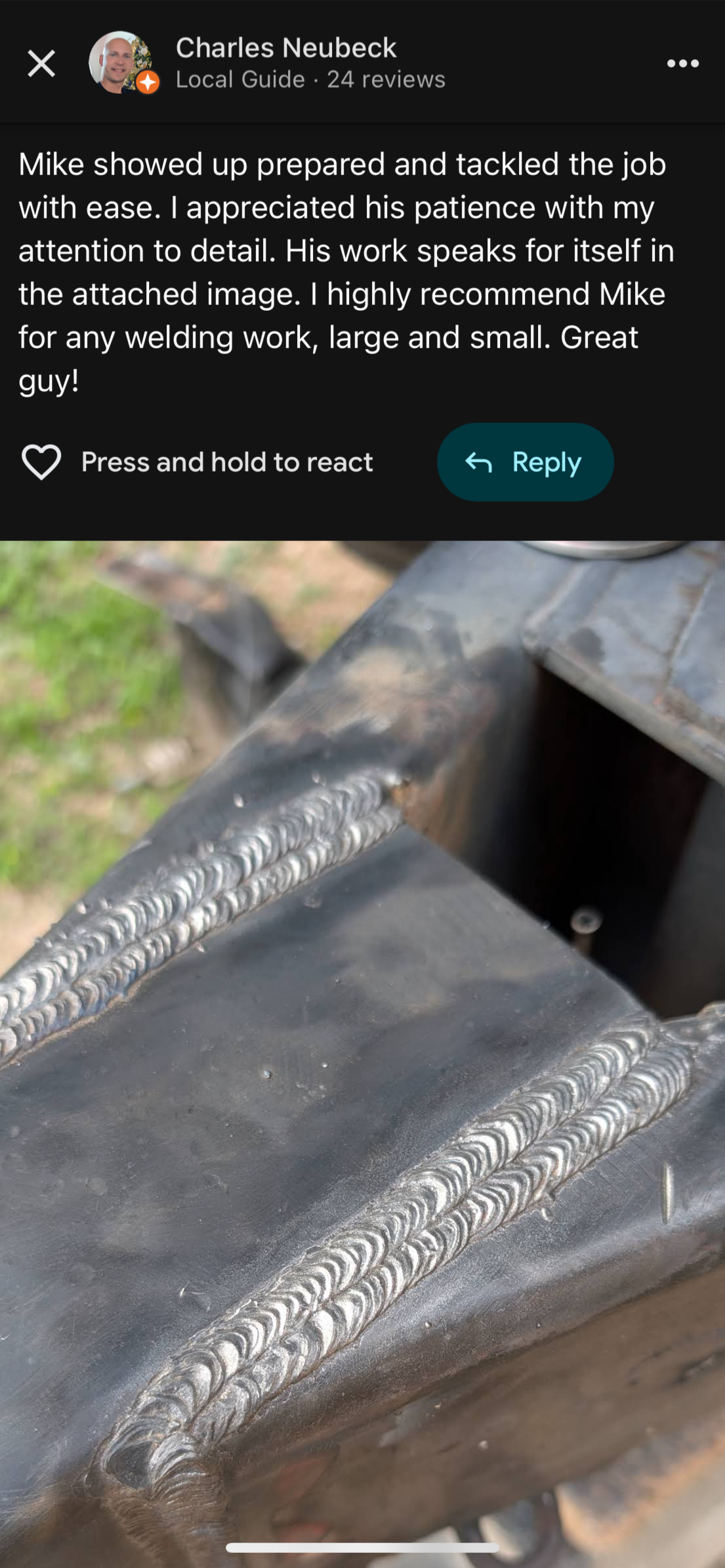 Close-up view of metal welding on steel, showing two bead welds along a joint, with a slightly blurred background of grass and other machinery parts.