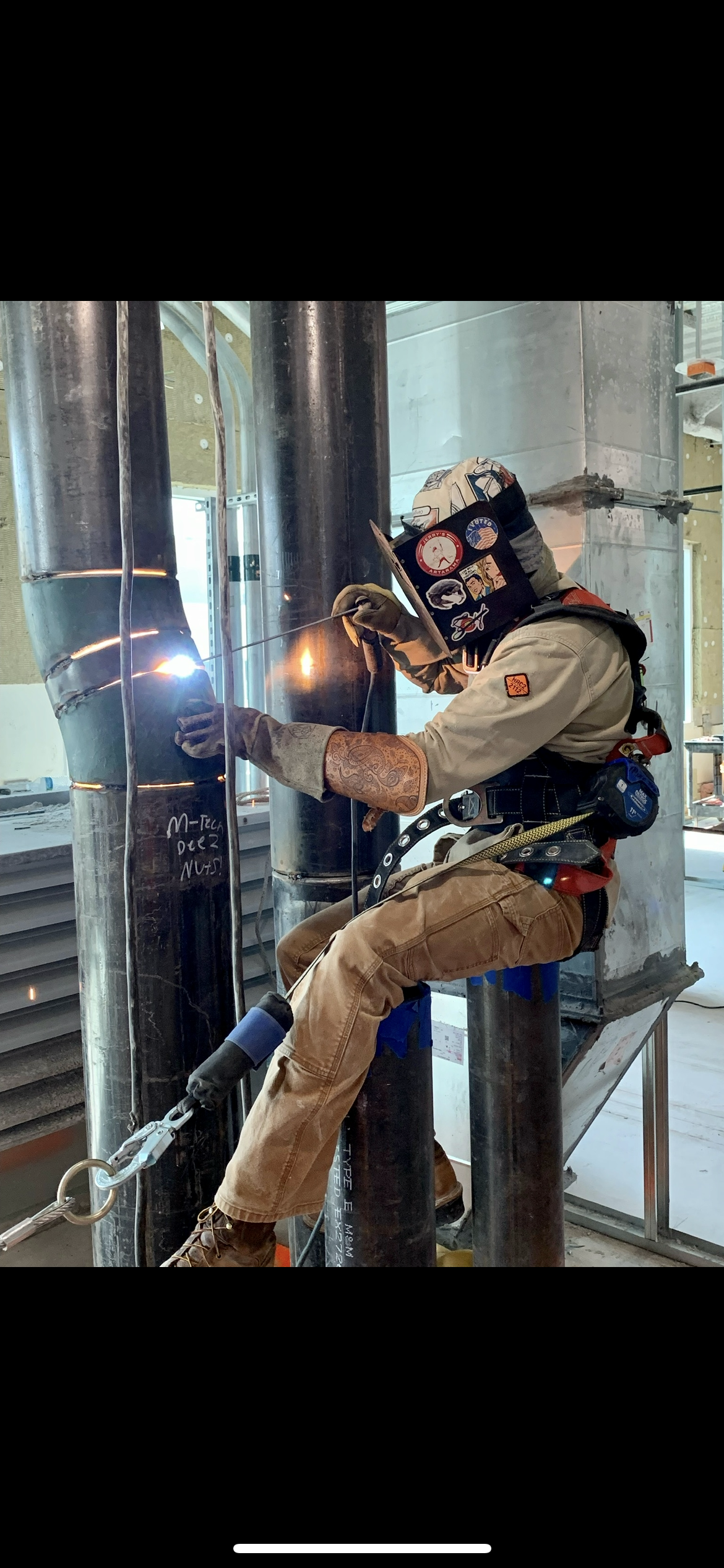 A worker performing welding on metal pipes inside a building. The worker is wearing protective gear including a welding helmet, gloves, and safety harness.