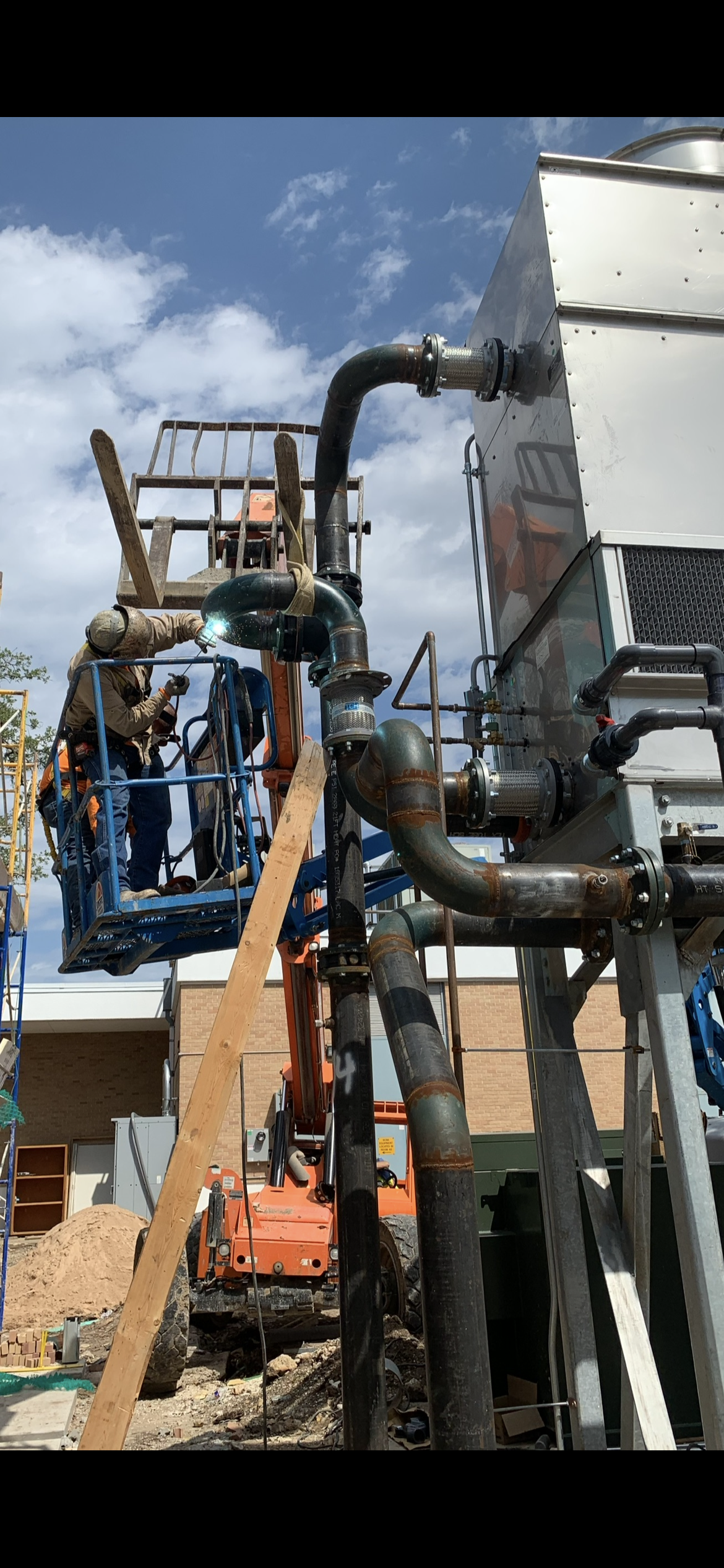 Construction worker welding pipes on a scaffolding next to a large industrial machine outdoors with a brick building and blue sky in the background.