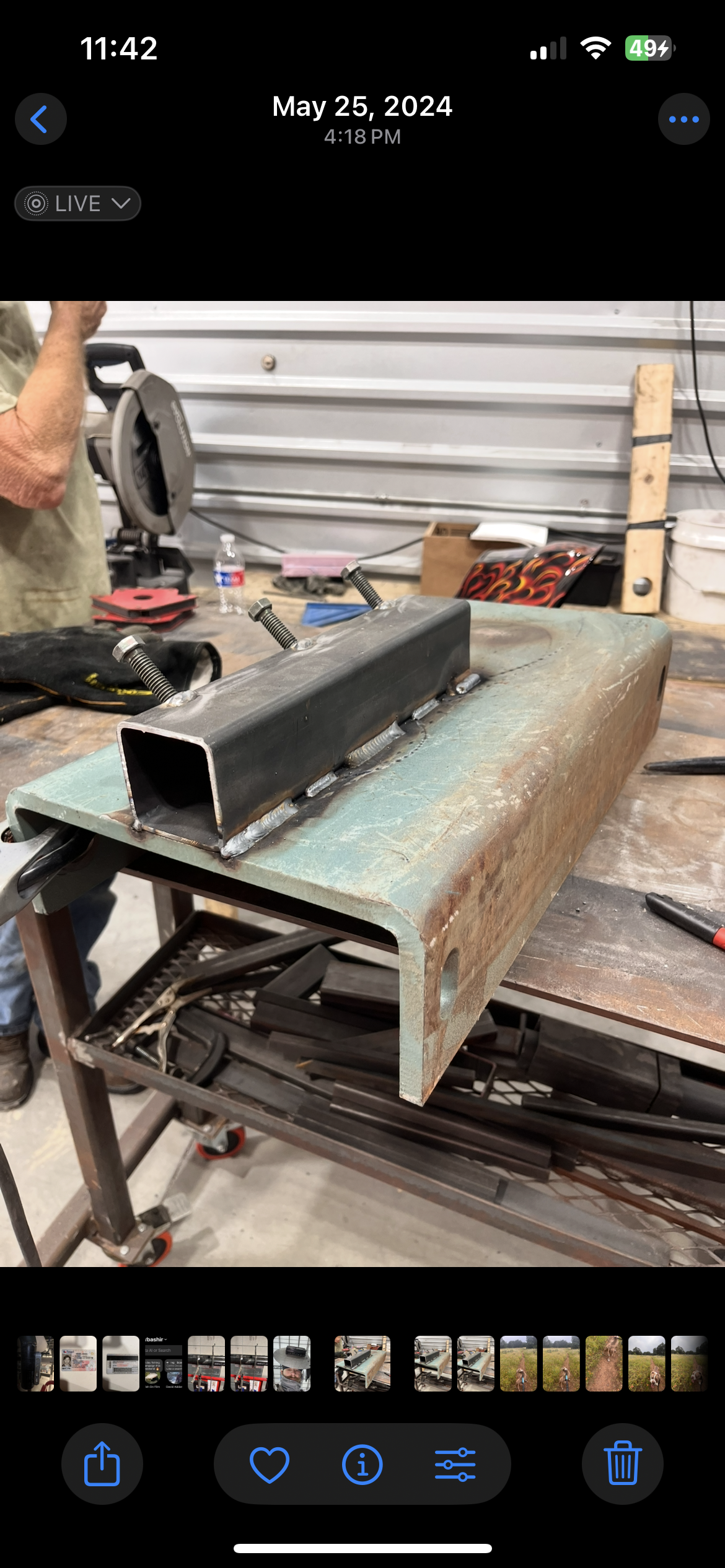 A metal rectangular tube welded onto a black metal base, with three bolts protruding from the top. The setup is on a workbench in a workshop, with tools, a water bottle, and a person holding a saw in the background.