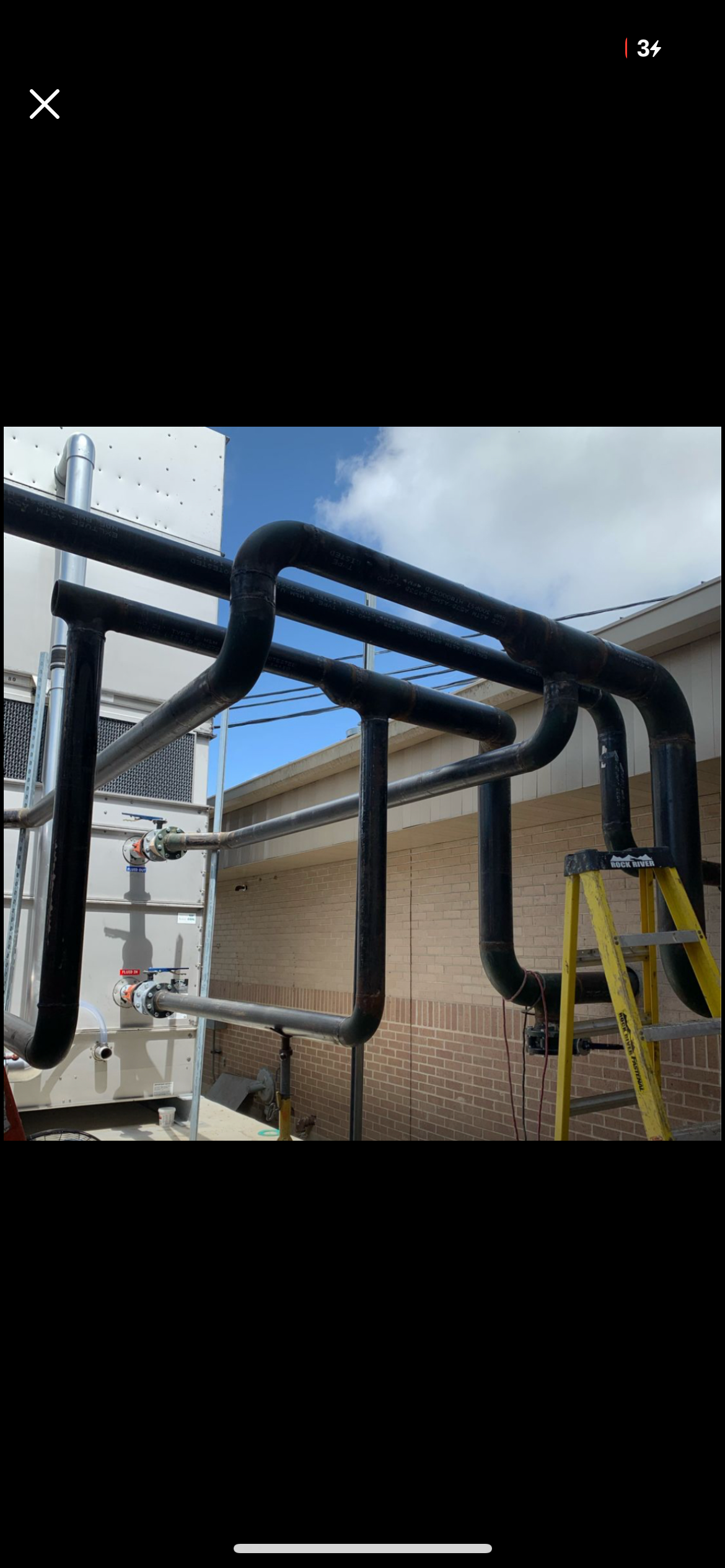Black pipes and a yellow ladder outside a building with a brick wall, part of a cooling or HVAC system, under a partly cloudy sky.