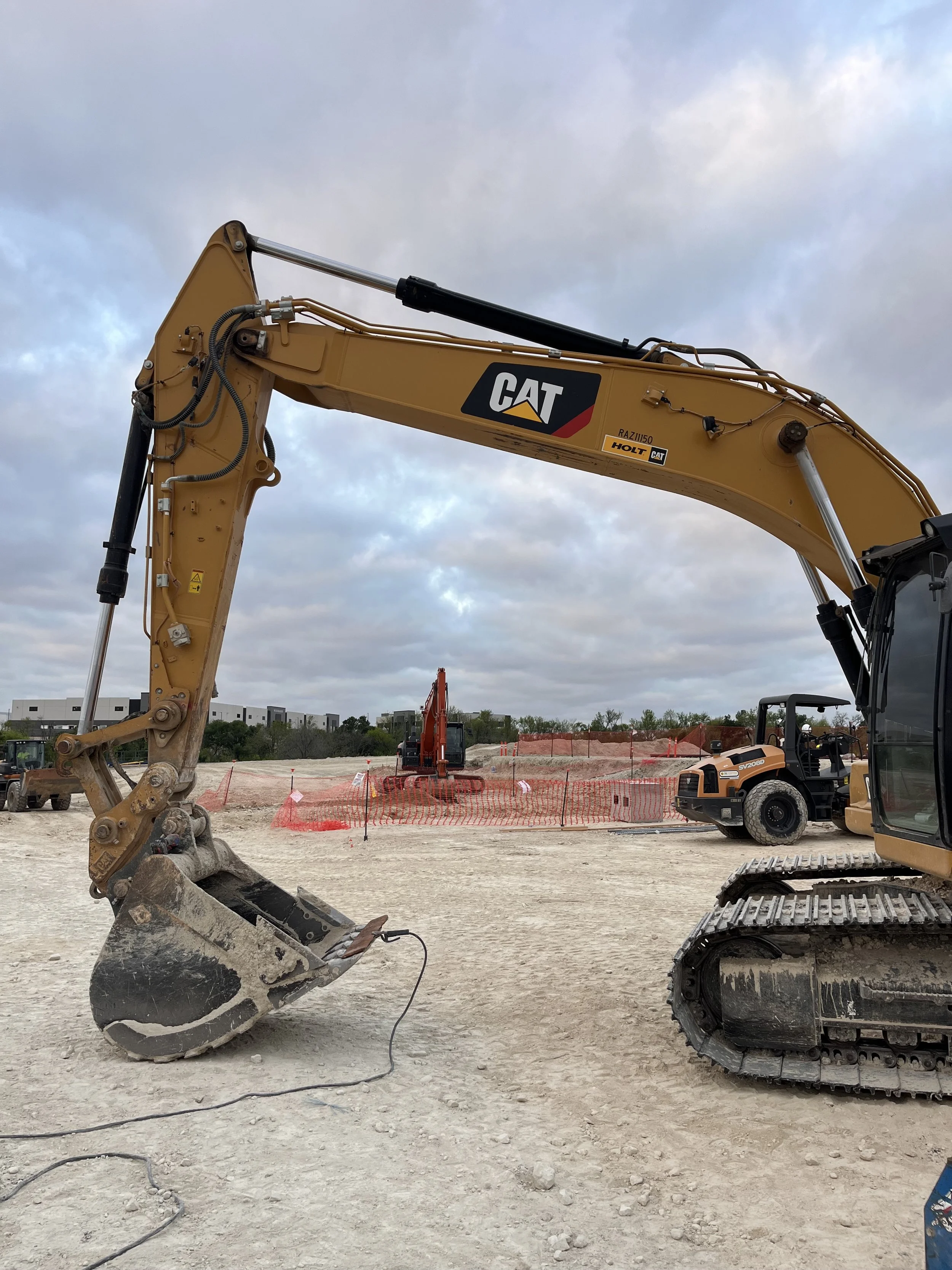 A construction site with a large yellow CAT excavator and tracked machinery on dirt ground, with orange safety fencing and some construction equipment in the background under cloudy skies.