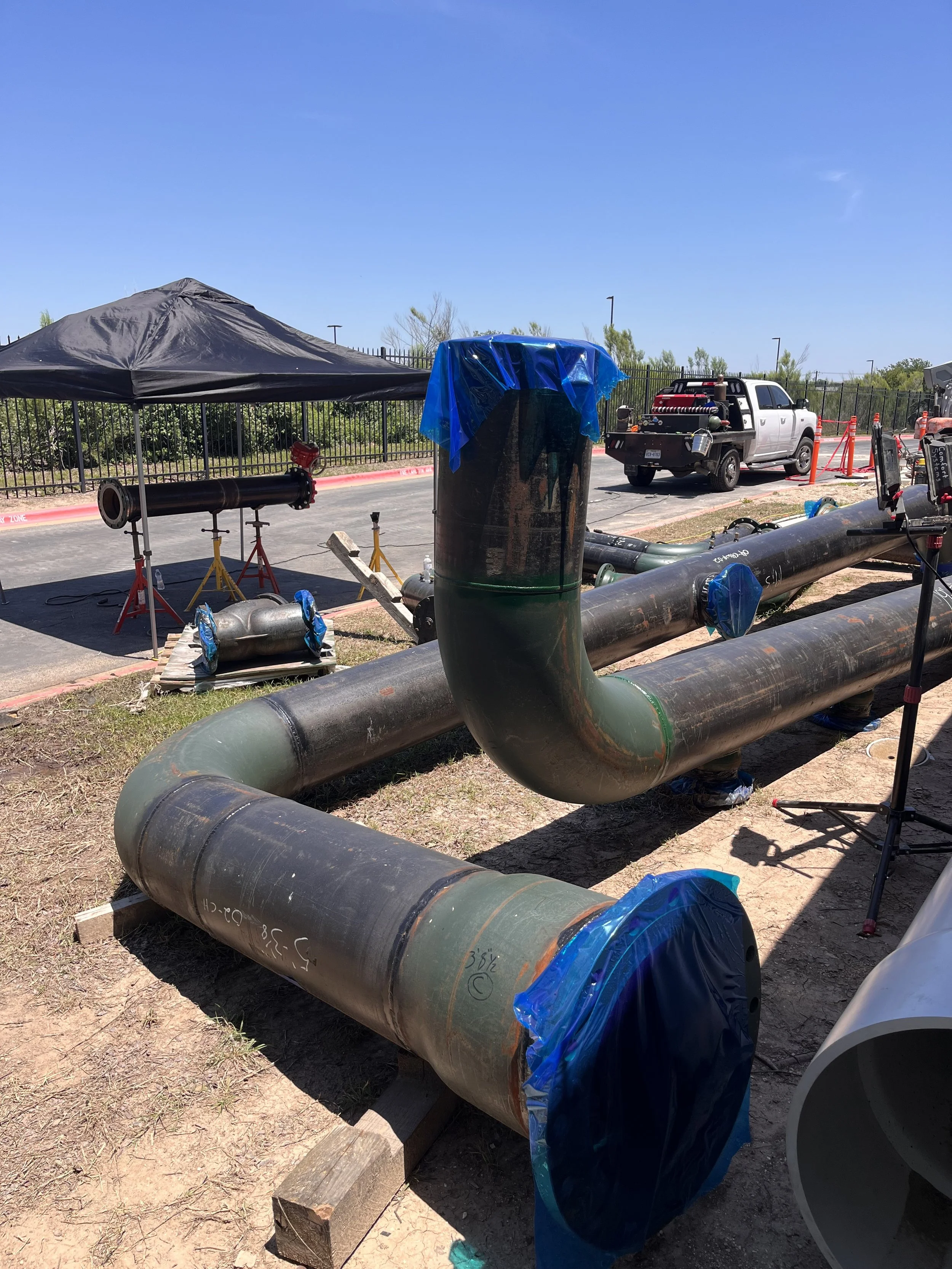 Outdoor construction or maintenance site with large black pipes, some covered with blue plastic, a portable work tent, a white work truck, and various equipment, outside on a sunny day.