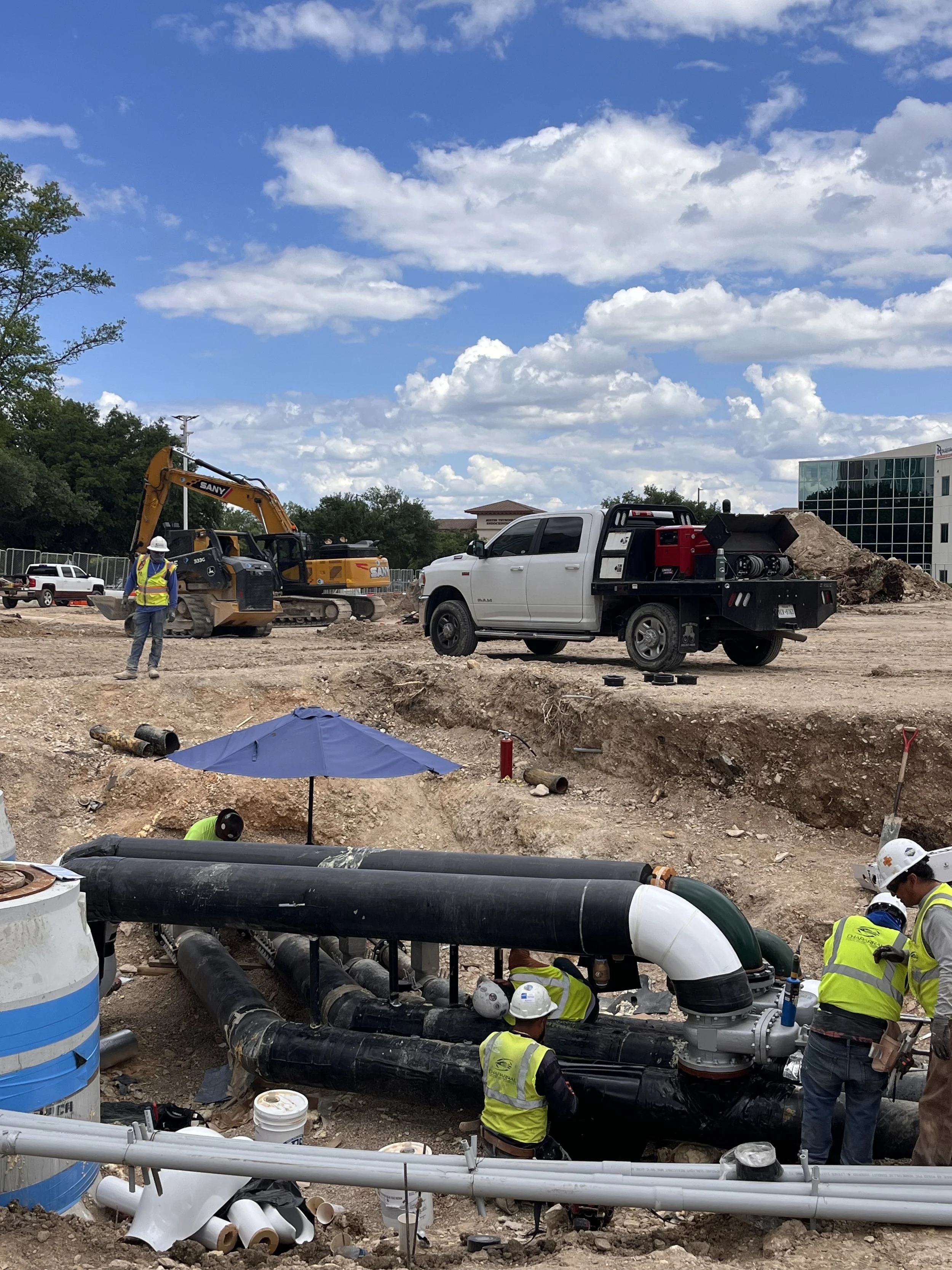 Construction workers in safety vests and helmets working on plumbing pipes at a construction site. Heavy machinery and large pipes are visible, with a partly cloudy sky above.