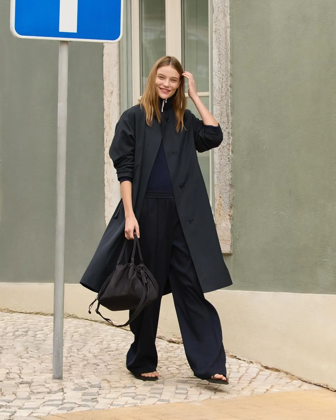 A young woman with long hair smiling, walking on a cobblestone sidewalk, wearing a black coat, black pants, and flip-flops, carrying a black bag.