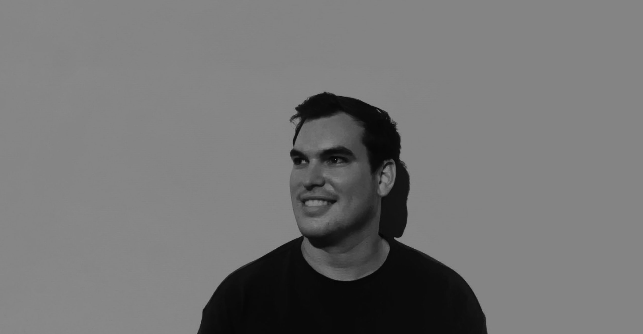 Black-and-white photo of a smiling man with dark hair, wearing a black shirt, looking slightly to the right against a plain background.
