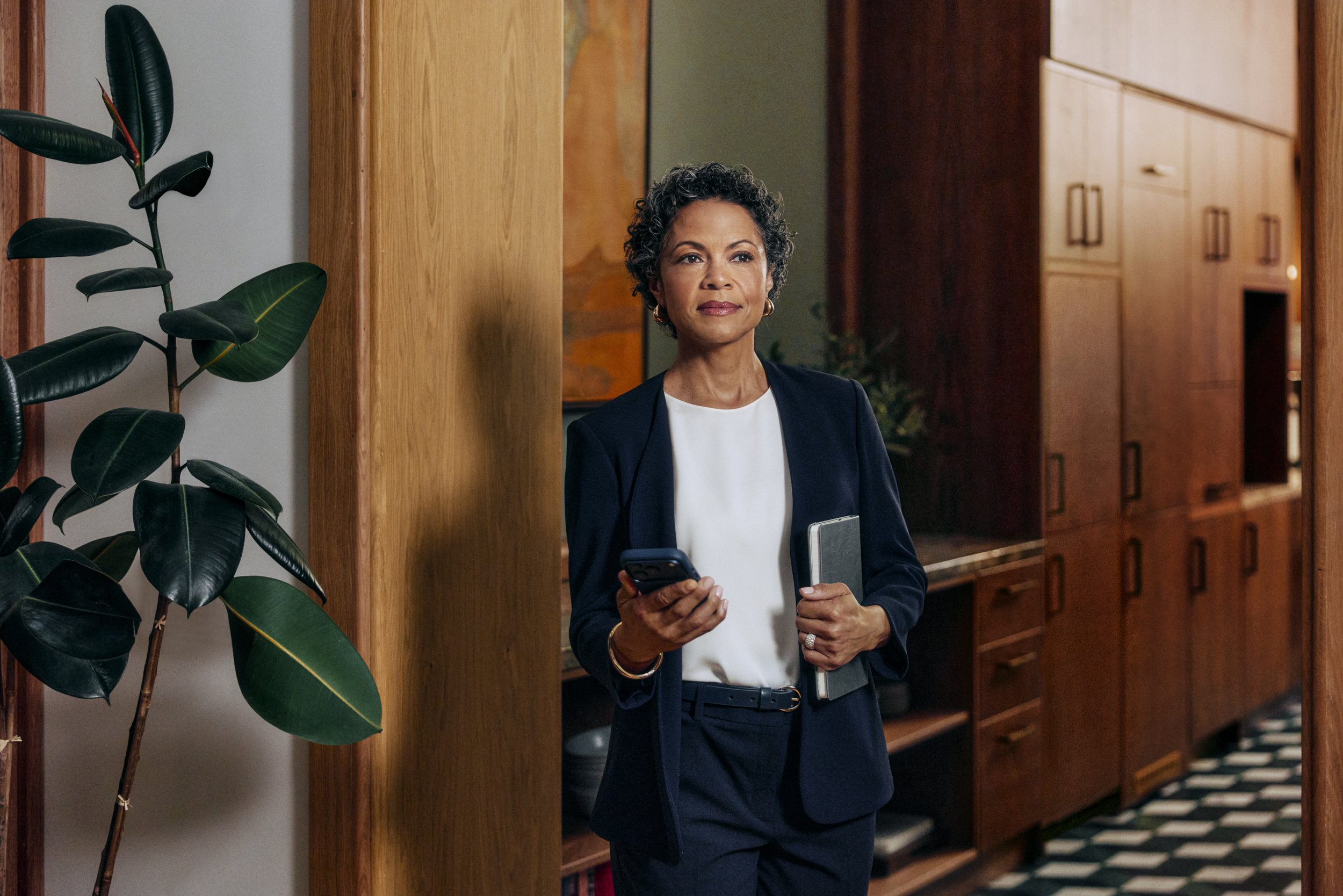 A woman with gray curly hair, dressed in a navy blazer and white top, holding a tablet and a smartphone, standing in what appears to be a library or office with wooden shelves and cabinets.