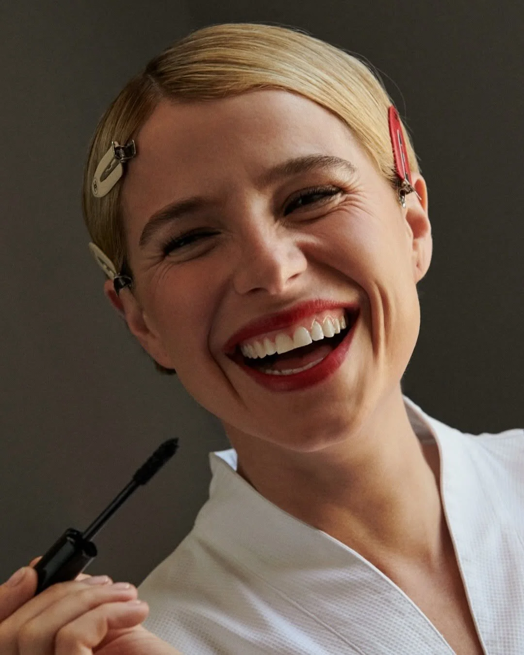 A woman smiling widely, with blond hair held back by hair clips, wearing red lipstick and a white top, holding a makeup brush.